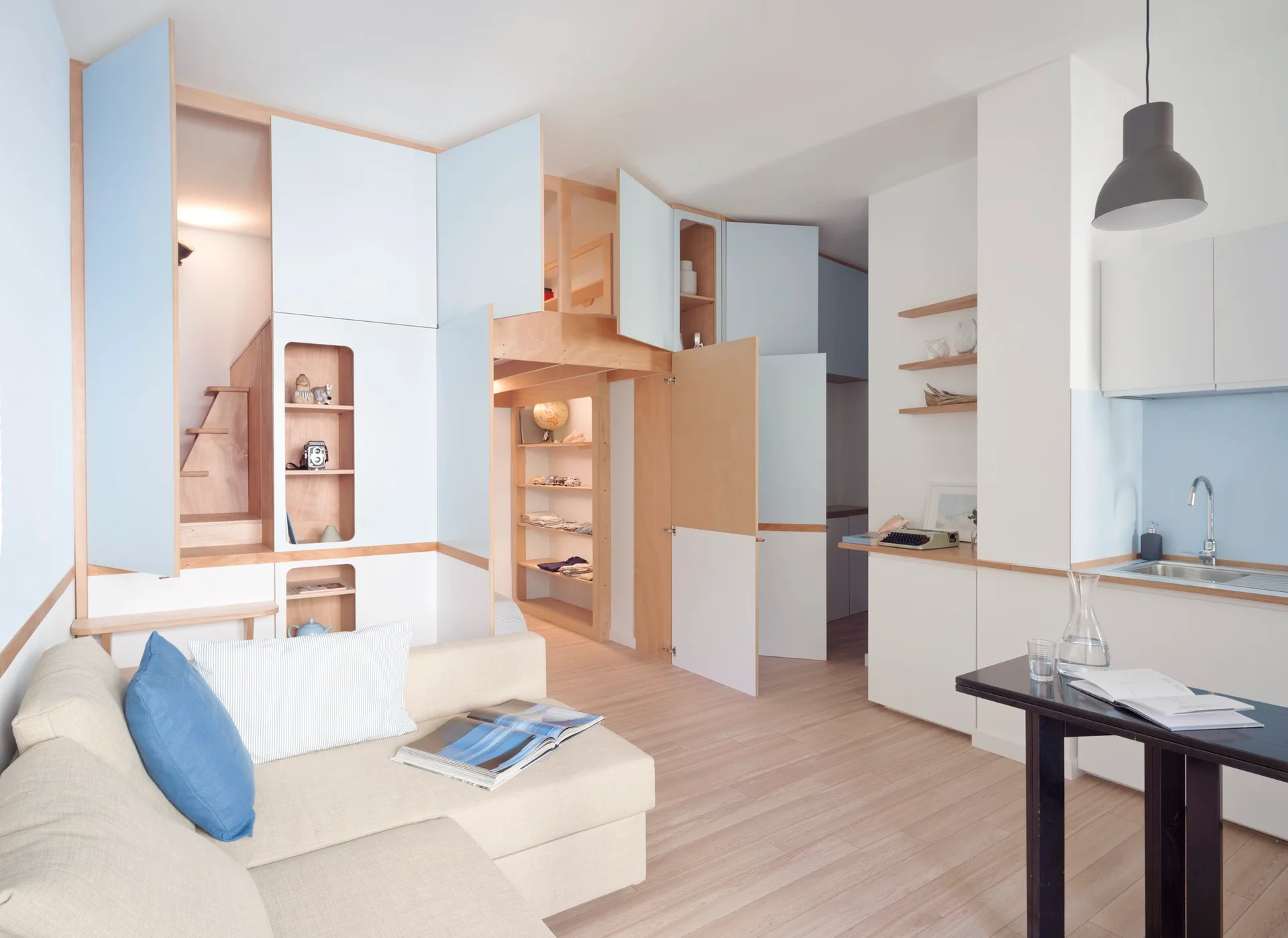 A compact micro-apartment interior features a cream sofa, a dark dining table, and a large pale blue and plywood storage unit with integrated loft stairs.