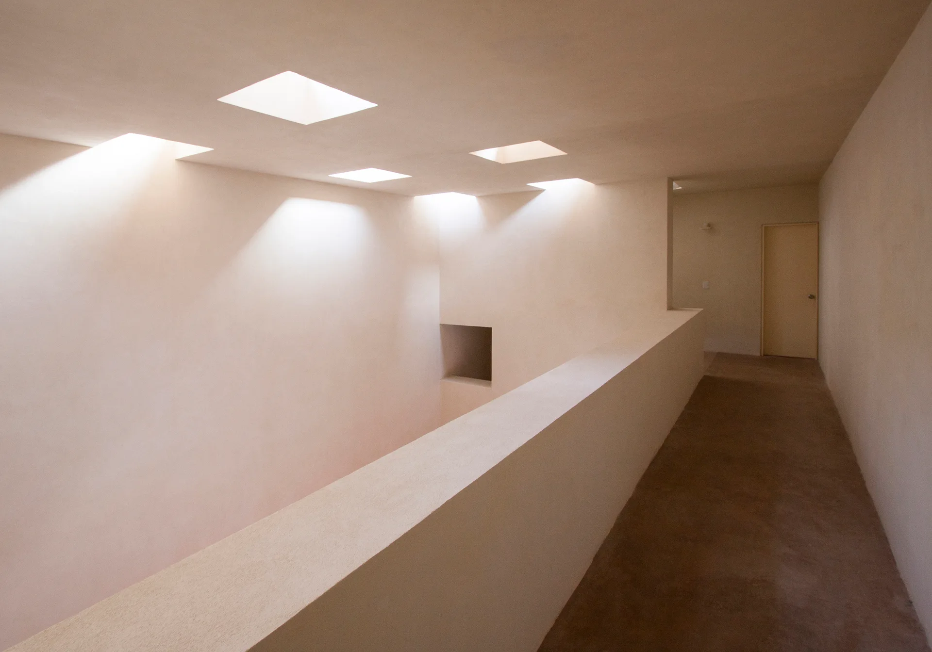 A minimalist hallway and landing area with smooth cream plaster walls and square skylight cutouts in the ceiling filtering natural light.