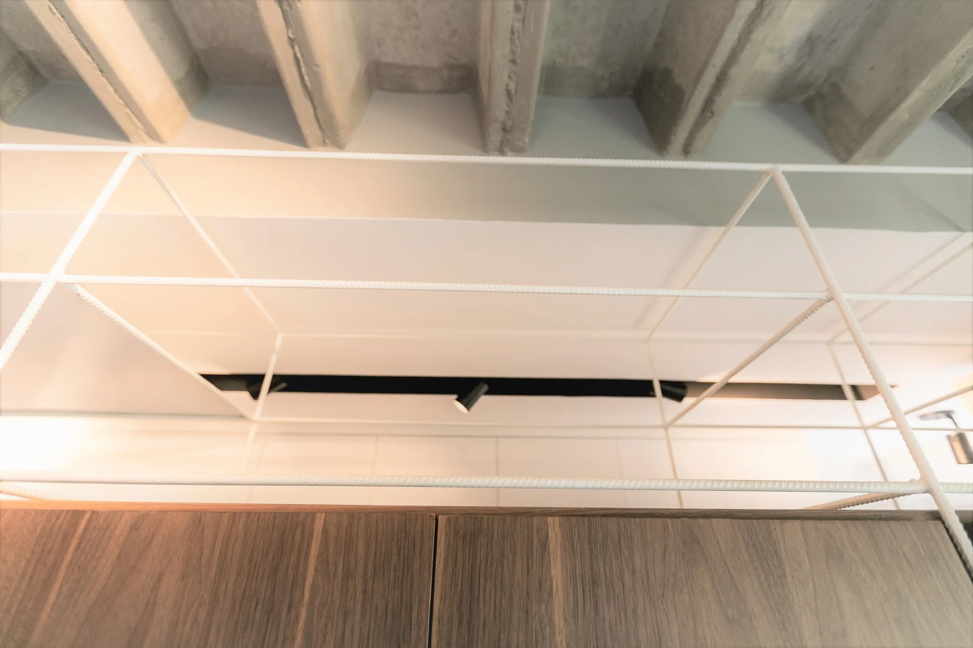 A low angle view of a white wire grid shelving frame suspended beneath rough concrete ceiling joists and above a wood cabinet.