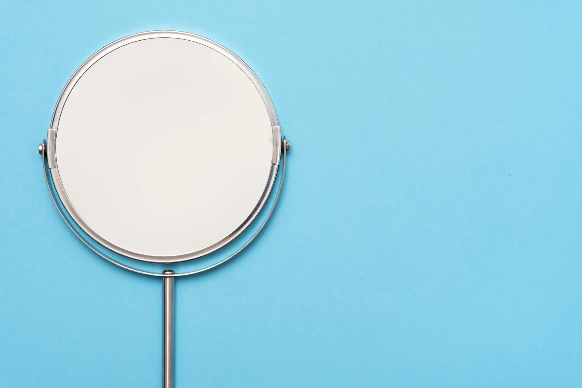 A round silver metal vanity mirror with a swivel stand set against a solid light blue background.