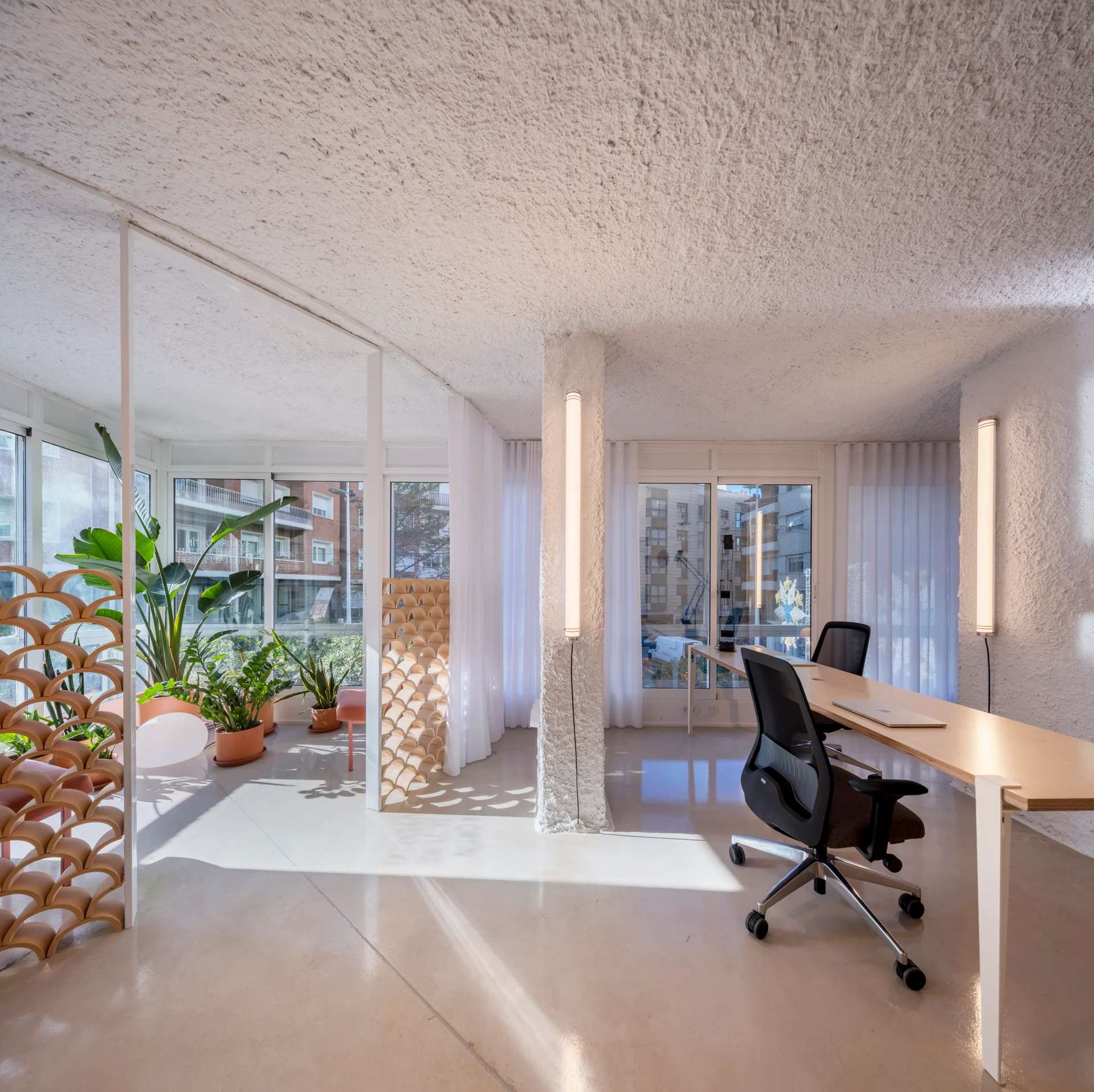 A minimalist office features a long wood desk, black office chairs, a heavily textured ceiling, and vertical tube lights mounted on a column.