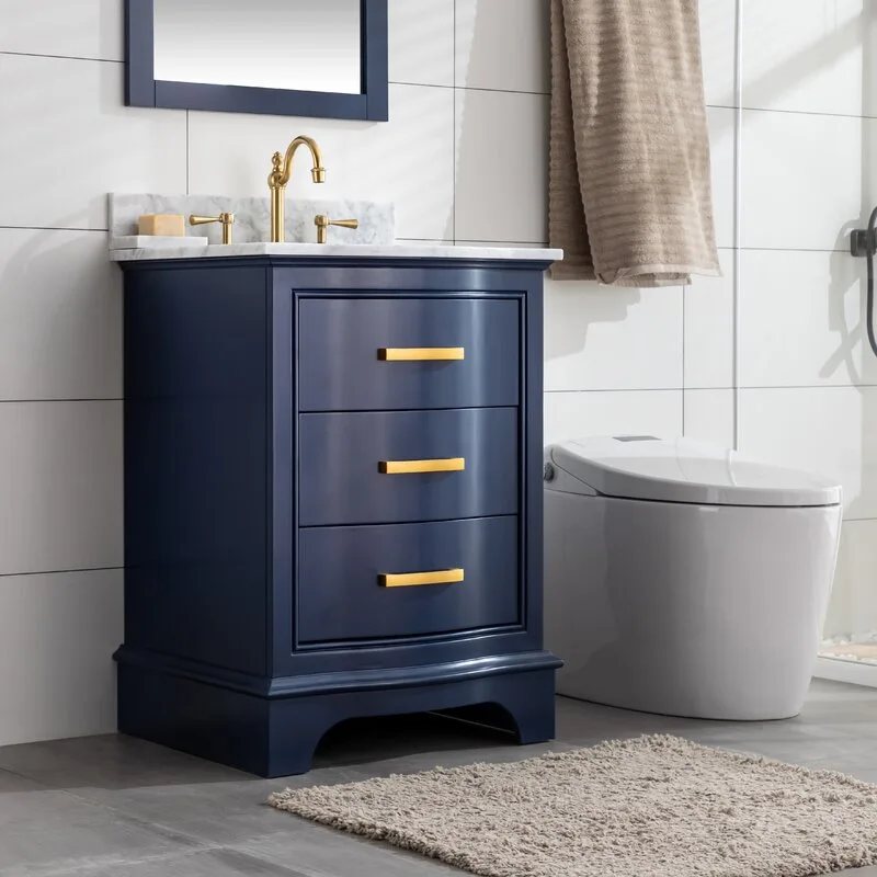 A modern navy blue bathroom vanity with a white marble top and gold faucet stands against a white tiled wall next to a toilet.