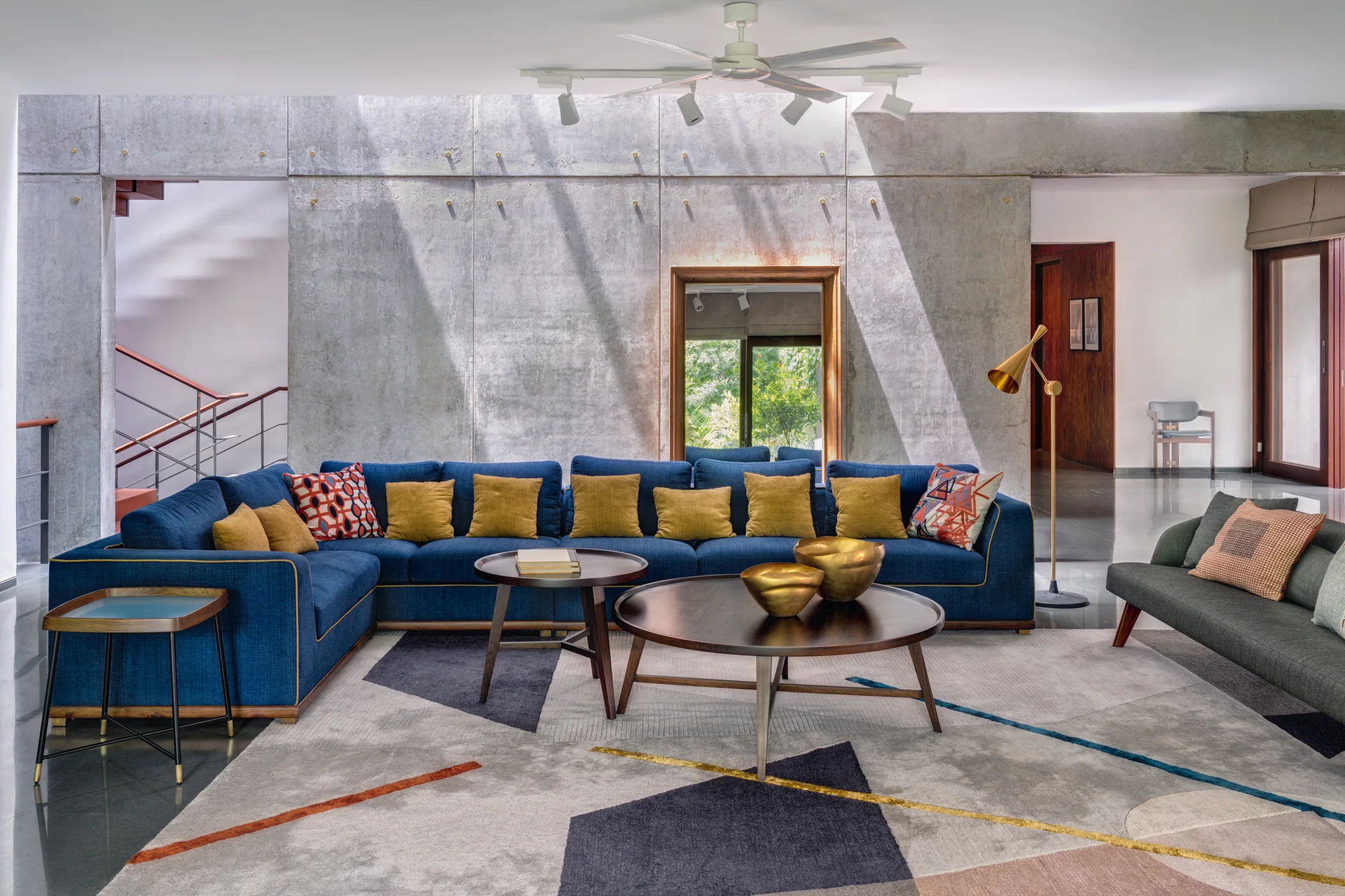A modern living room featuring a large blue sectional sofa, wooden coffee tables, and a geometric rug set against a textured concrete panel feature wall.