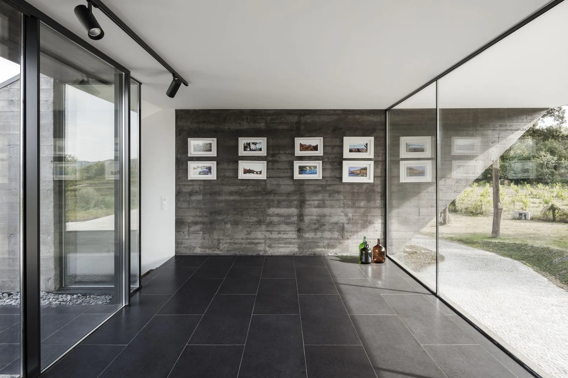 A minimalist room features a textured grey concrete wall with eight framed photos, dark tiled flooring, and expansive floor-to-ceiling glass windows.