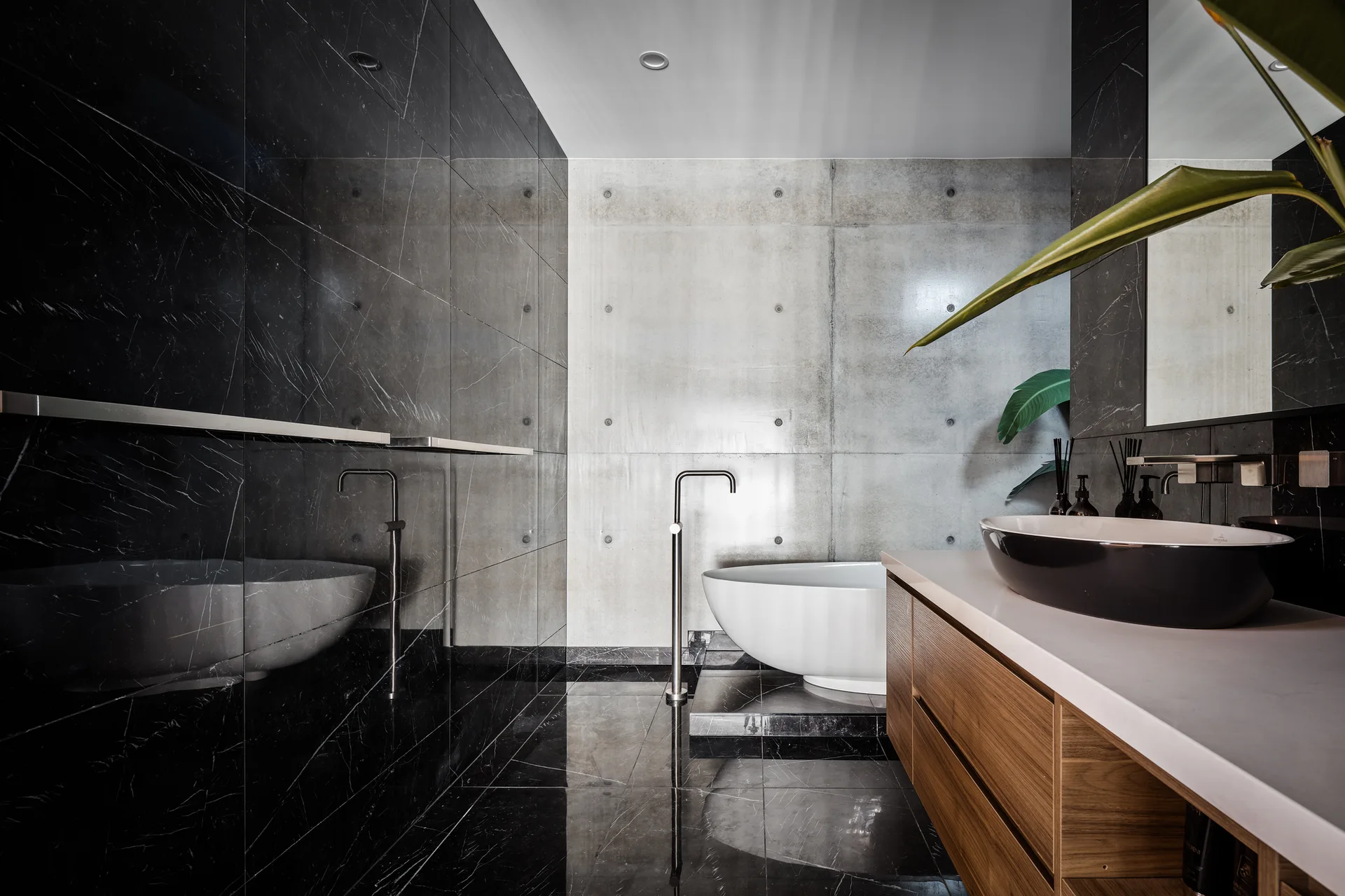 A minimalist bathroom with a white freestanding bathtub against a grey concrete wall, adjacent to a glossy black marble tiled wall and a wooden vanity.