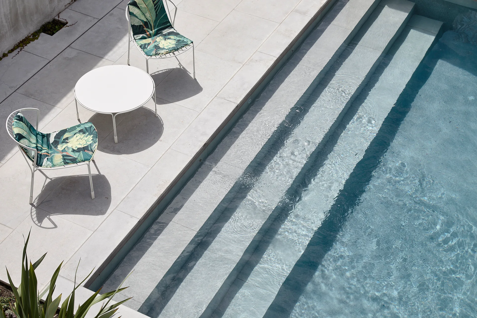 An overhead view of a light stone pool deck next to blue water, featuring two white metal chairs with green palm leaf cushions and a round table.