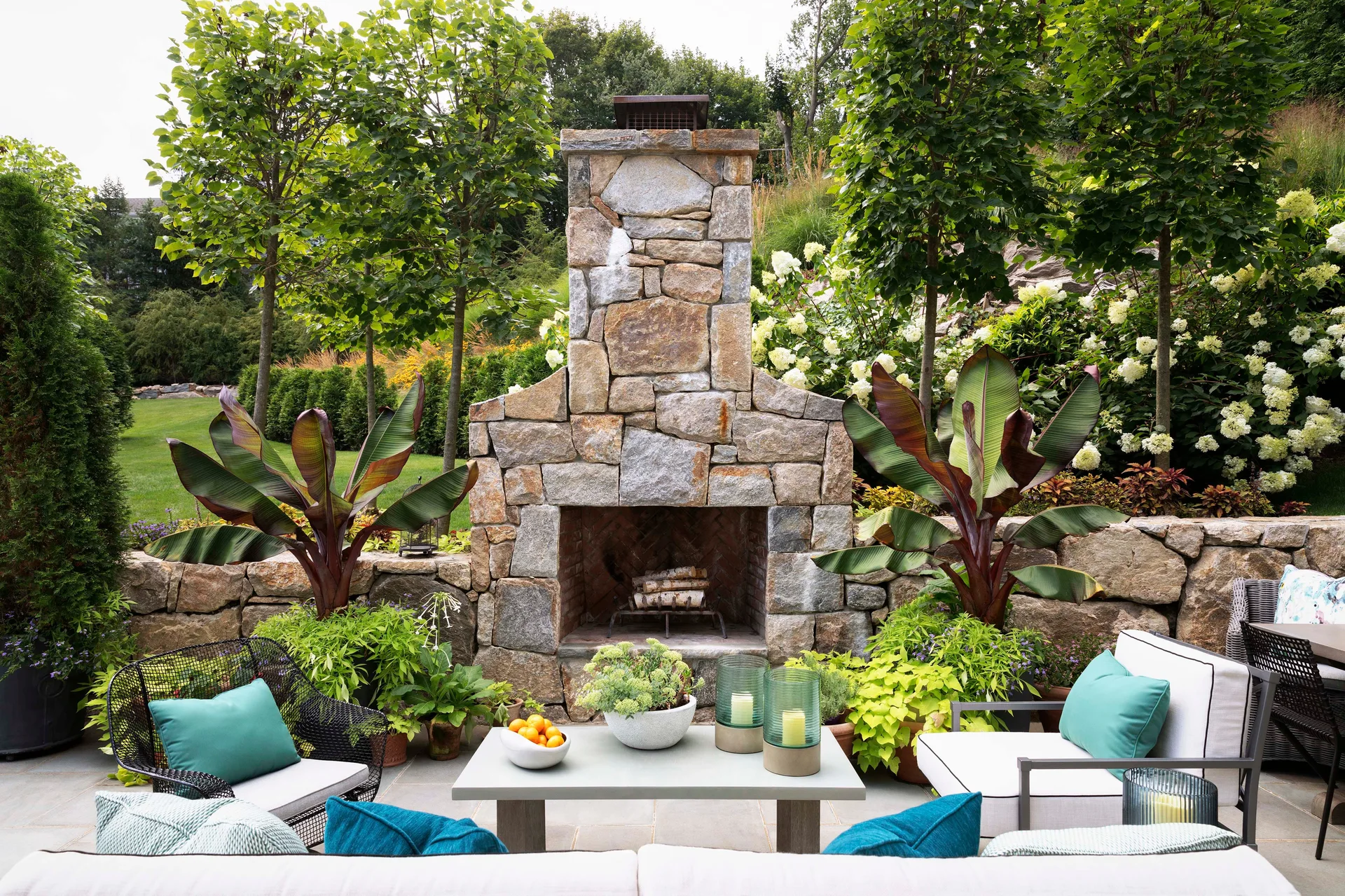An outdoor stone patio features a central fireplace, neutral seating with teal pillows, a coffee table, and potted plants against a lush green garden.