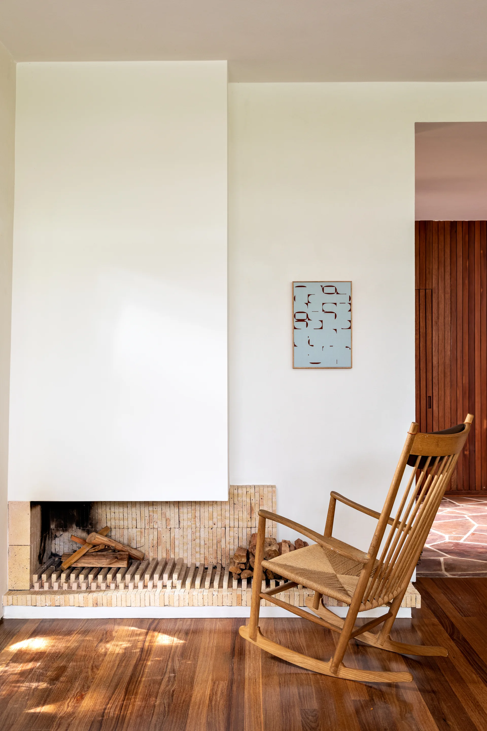 A wooden rocking chair with a woven seat sits on polished wood floors next to a minimalist white fireplace with a tan brick base.