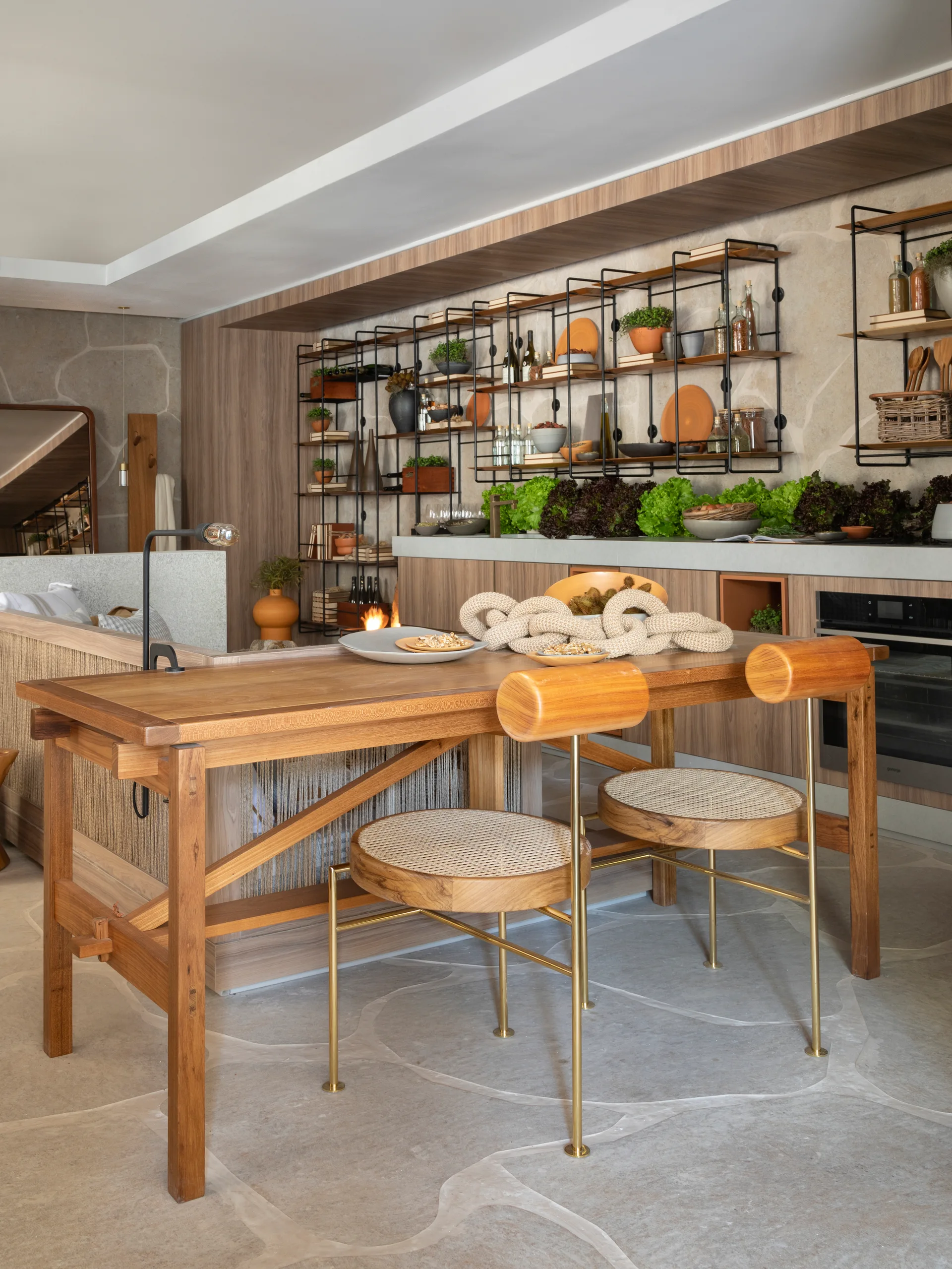 A contemporary kitchen features a wooden dining table with round cane-seated chairs in front of black metal open shelving and stone-textured walls.