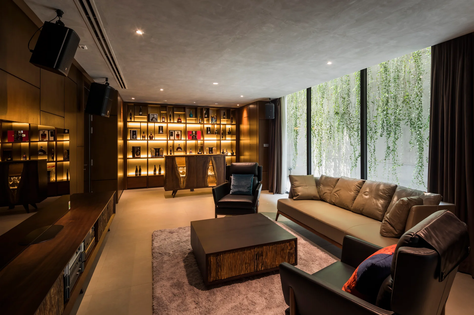 A contemporary living room featuring a leather sofa, dark wood coffee table, backlit display shelving, and a large window facing hanging green vines.