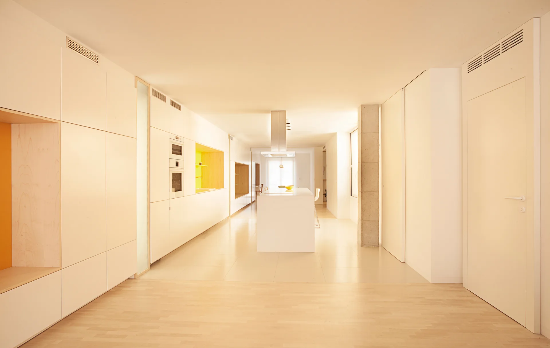 A minimalist white kitchen features handleless floor-to-ceiling cabinets, a central island, and a structural concrete column next to light wood floors.