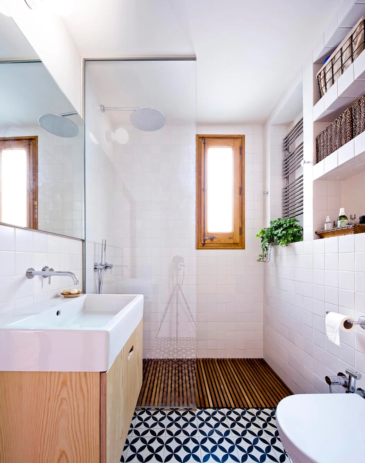 A modern bathroom features a light wood vanity, white square wall tiles, a glass shower partition with a wooden floor grate, and patterned floor tile.