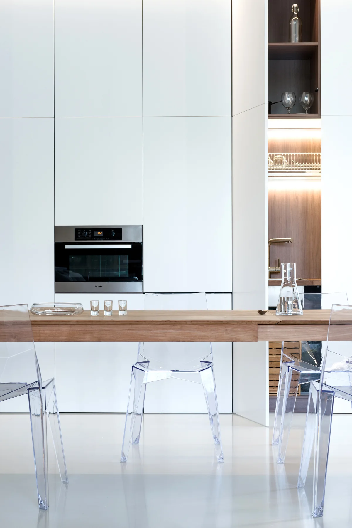 A light wood dining table with transparent acrylic chairs stands in front of minimalist white flat-panel kitchen cabinetry with a built-in oven.