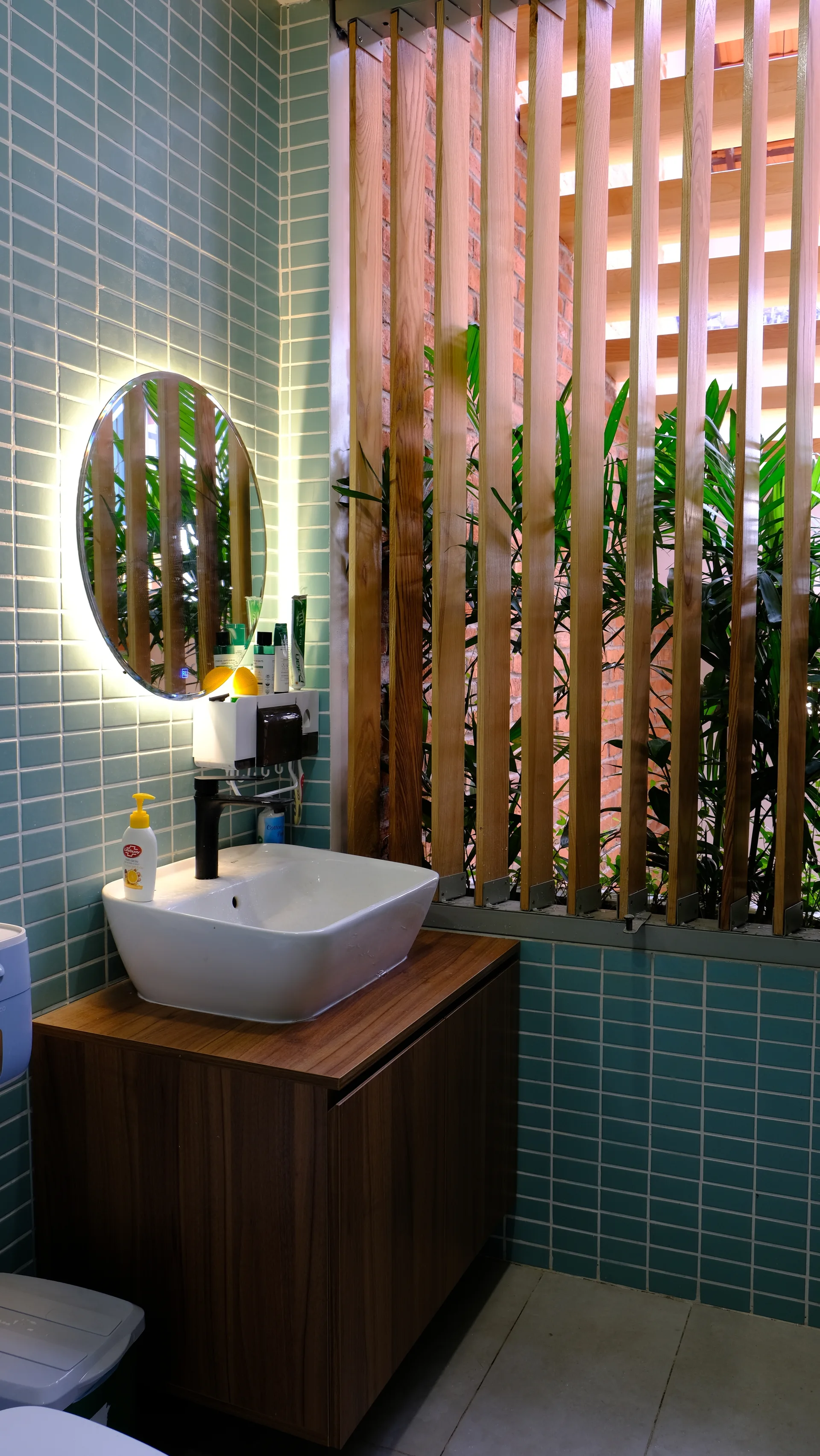 A modern bathroom features a white vessel sink on a wood vanity, a backlit round mirror against teal tiled walls, and a wooden slat partition.