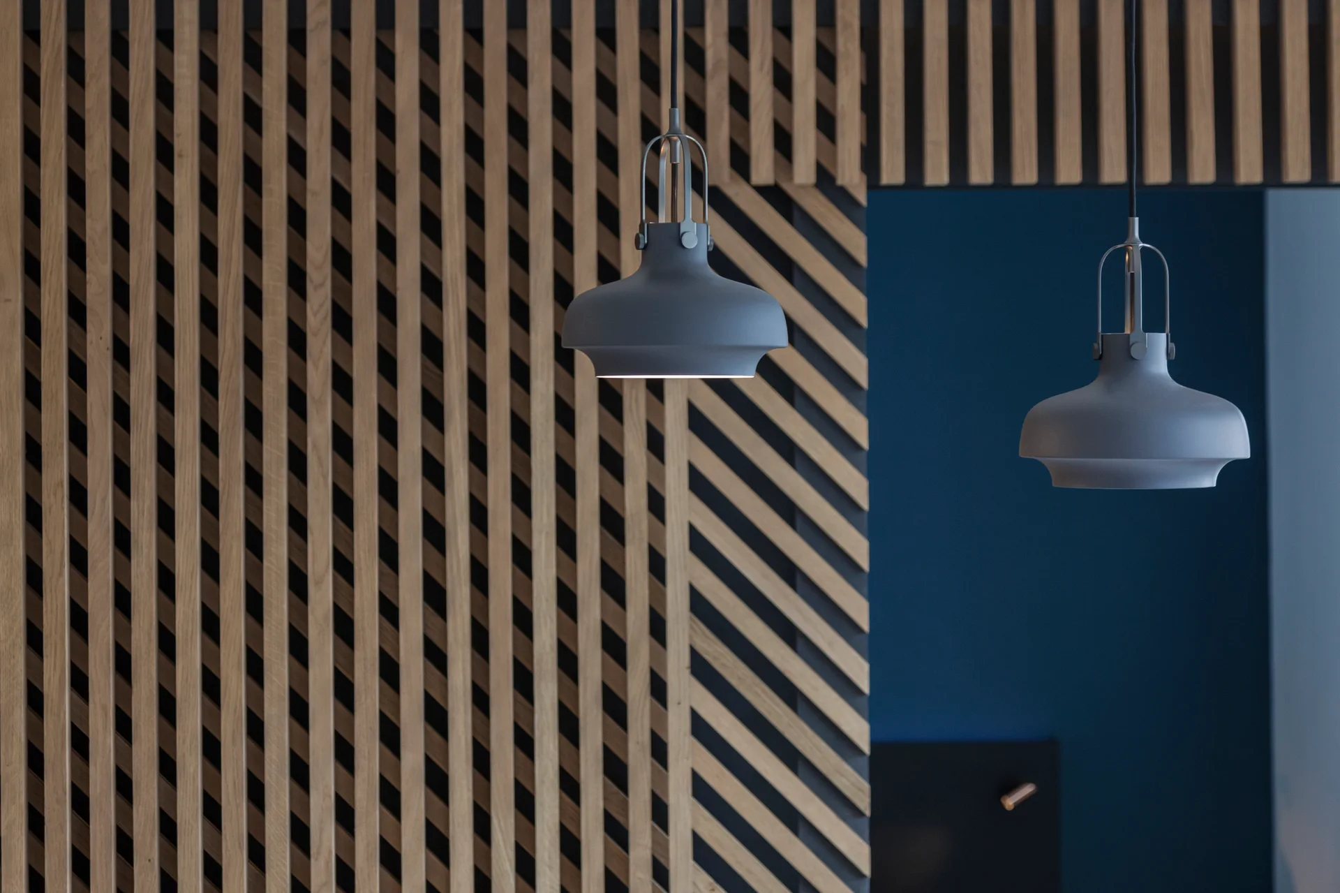 Two matte gray pendant lights hang in front of a modern wall featuring vertical and diagonal light wood slats over a deep blue background.