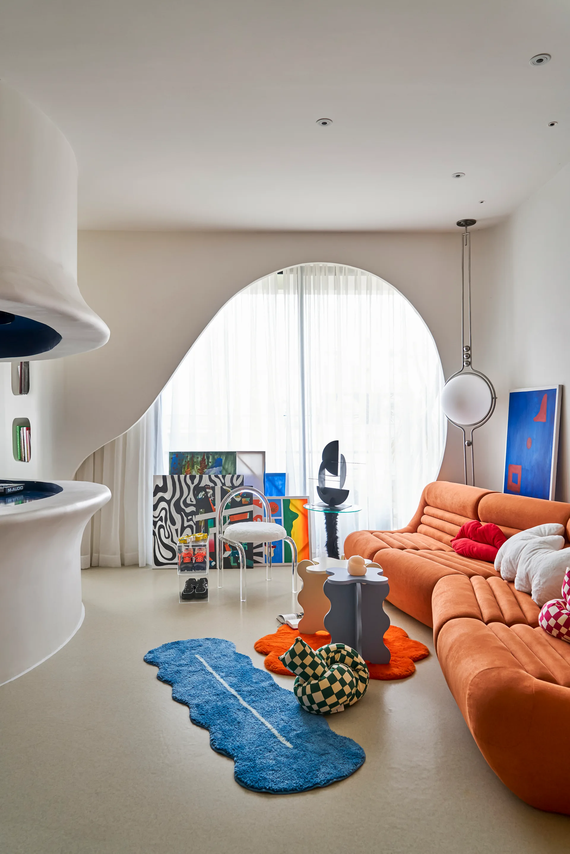 A bright living room featuring a curved orange sofa, an organic white plaster fireplace, and a large arched window with sheer curtains.