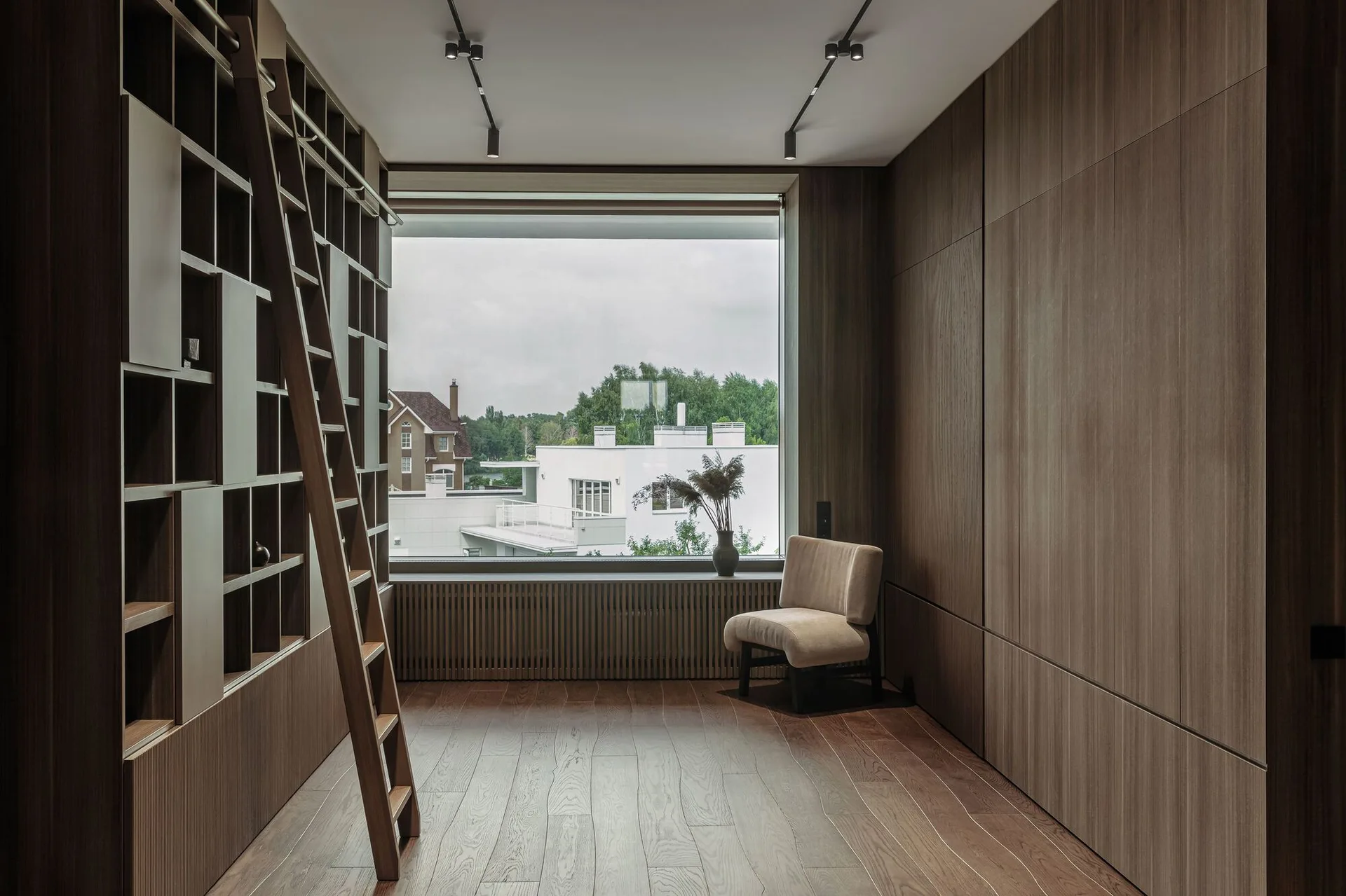 A minimalist home library features built-in wood bookshelves with a library ladder, a wood-paneled wall, and a lounge chair by a large window.