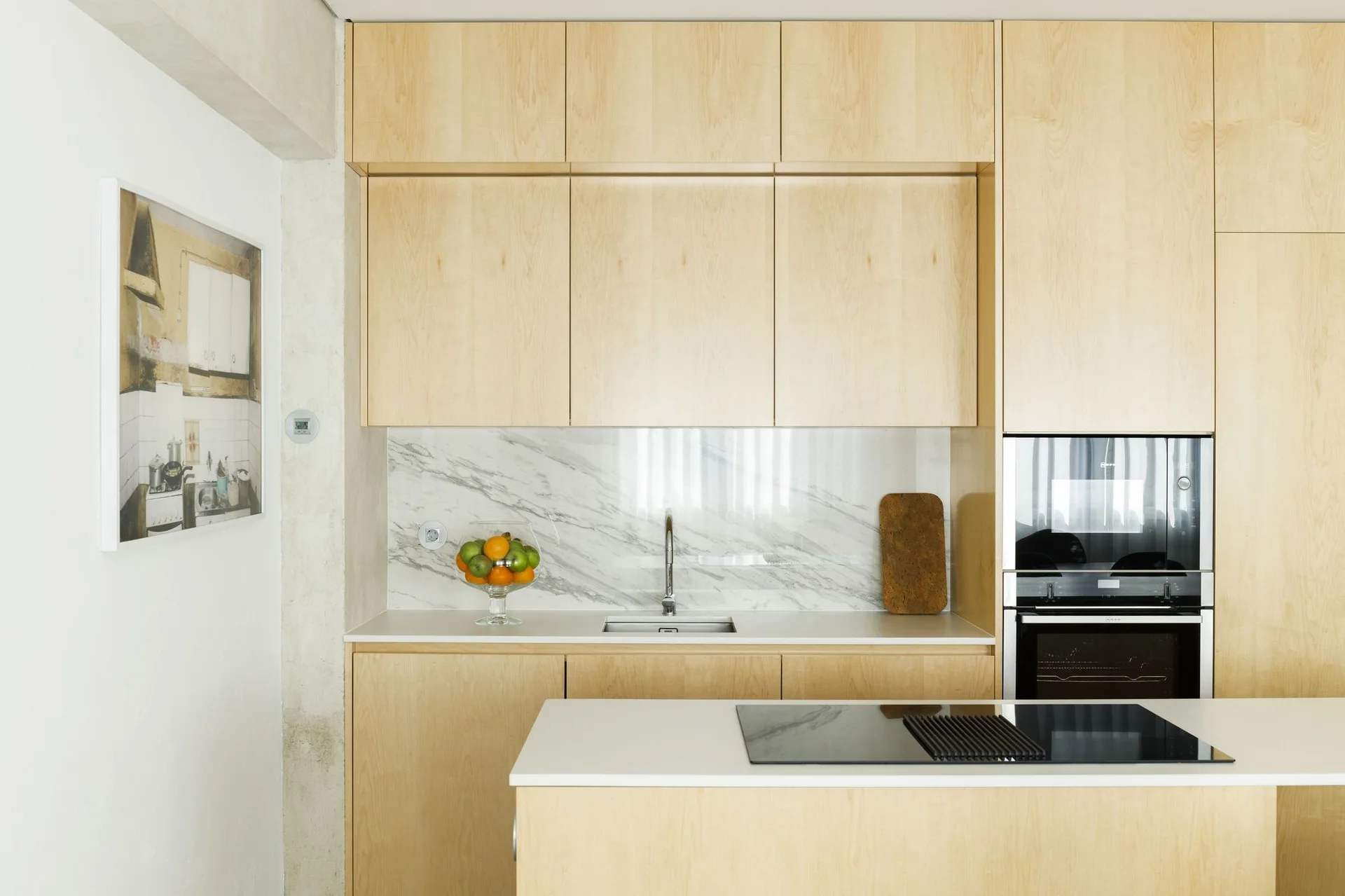 A modern kitchen featuring flat-panel light wood cabinetry, white countertops, an undermount sink, and a veined marble backsplash.