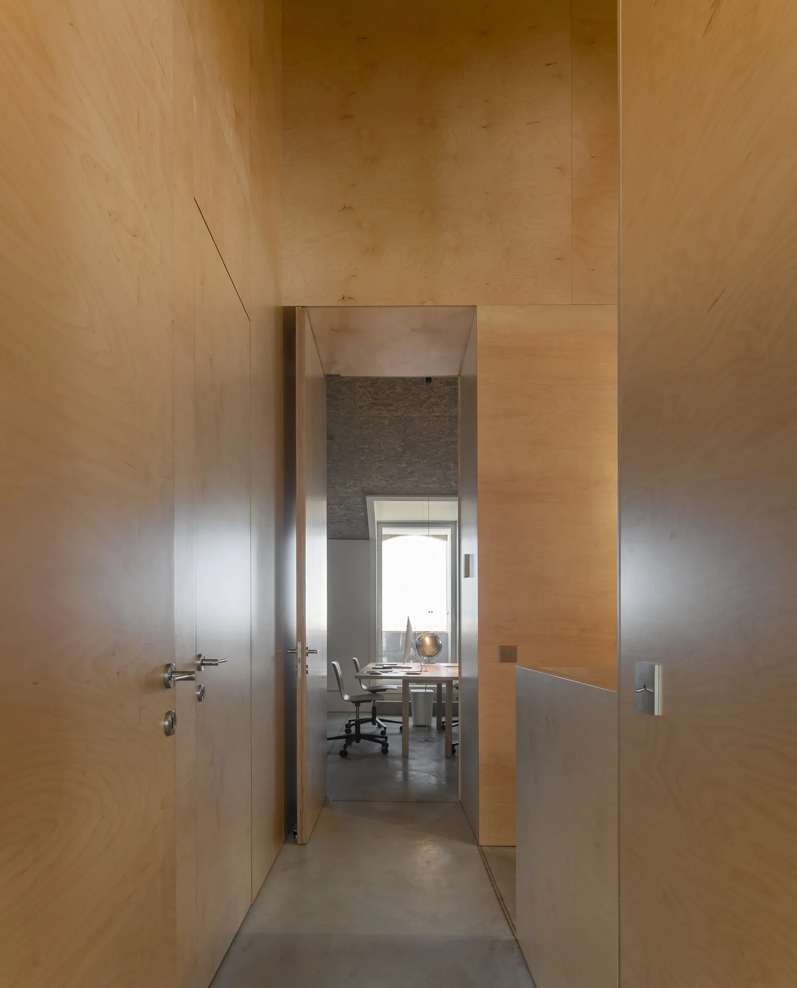 A narrow hallway with light wood plywood paneled walls and a concrete floor leads to a bright home office with a desk and chair.