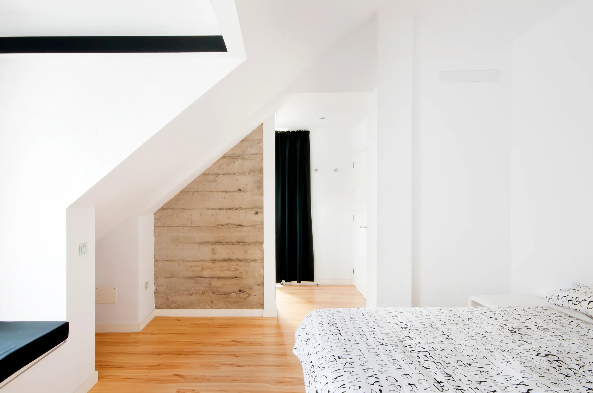 A minimalist attic bedroom features a bed with alphabet-print sheets, light wood floors, an angled ceiling, and an exposed concrete wall section.