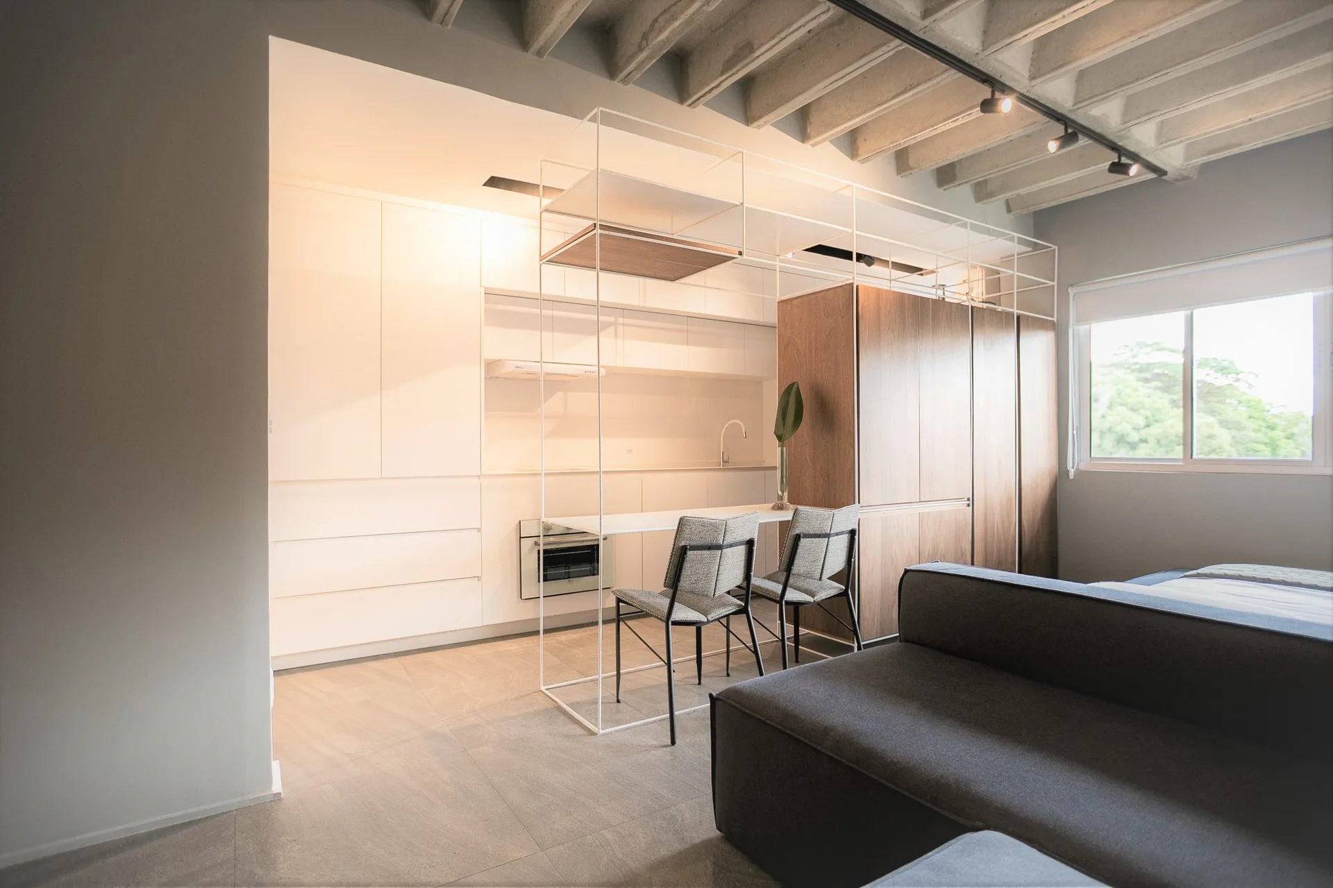 A minimalist kitchen features sleek white and wood-grain cabinets, an integrated metal frame dining island, and an exposed concrete beam ceiling.