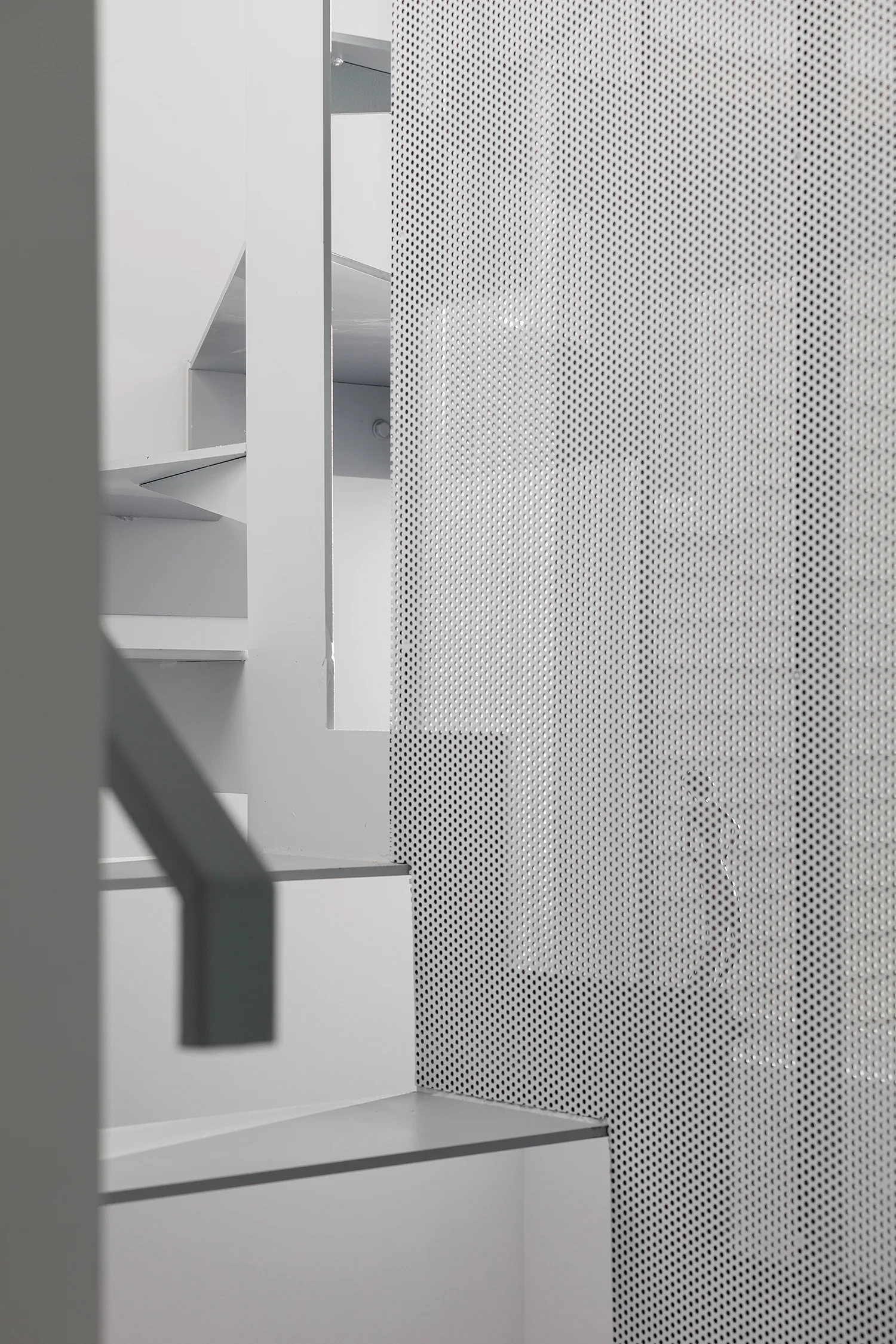 A close-up view of a minimalist white staircase featuring grey-edged treads, a geometric handrail, and a perforated metal safety screen.