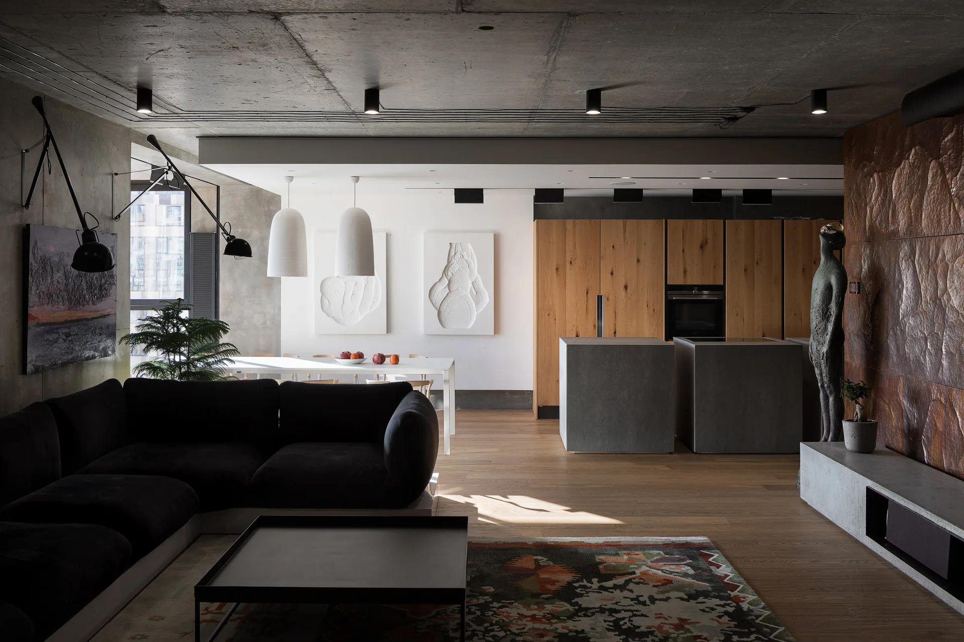 An industrial open-plan living room features a dark sectional sofa, a raw concrete ceiling with track lighting, and wood kitchen cabinetry.