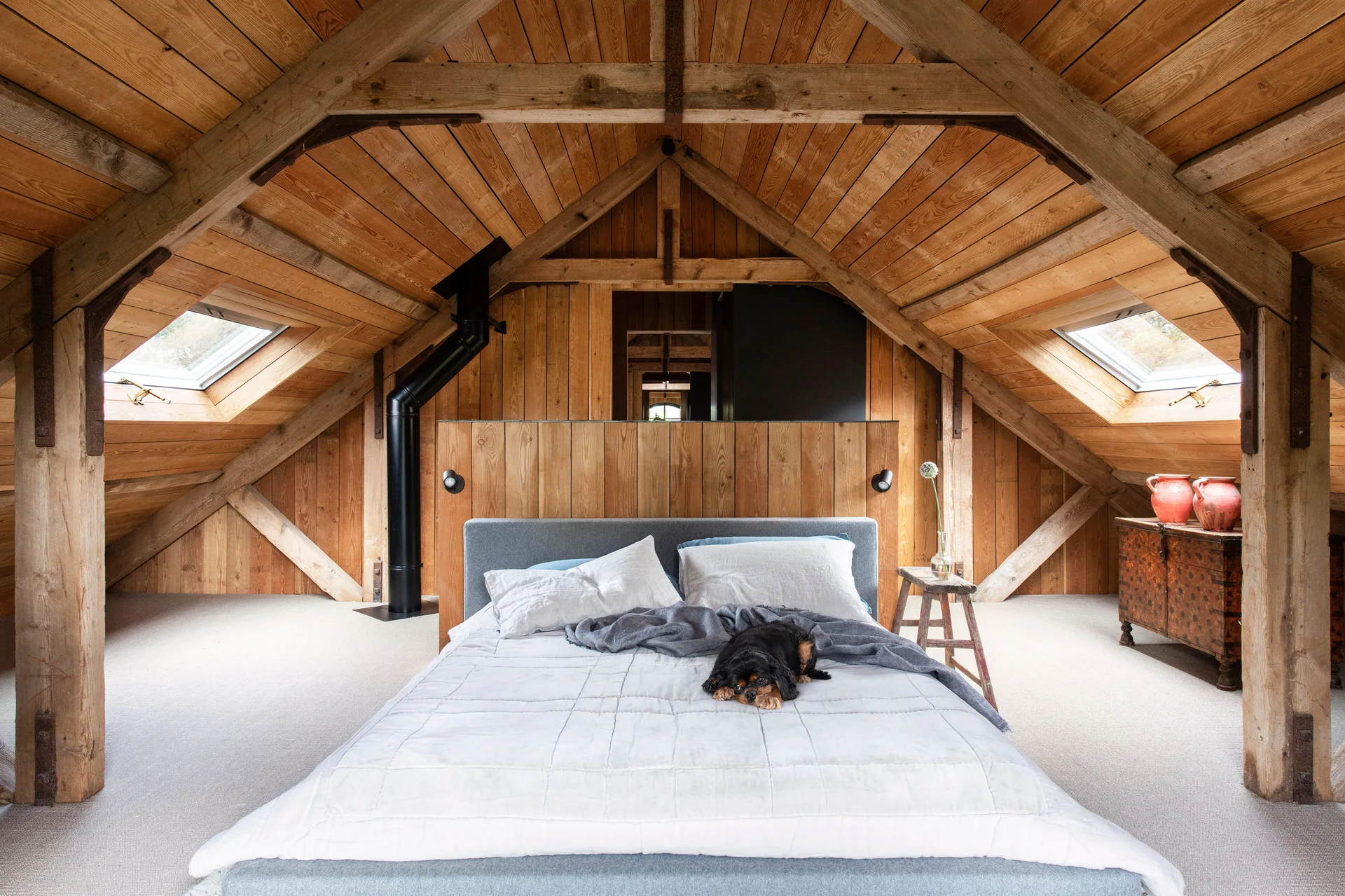 A rustic attic bedroom features a sloped wood-paneled ceiling with heavy timber beams, skylights, and a central bed with a gray headboard.