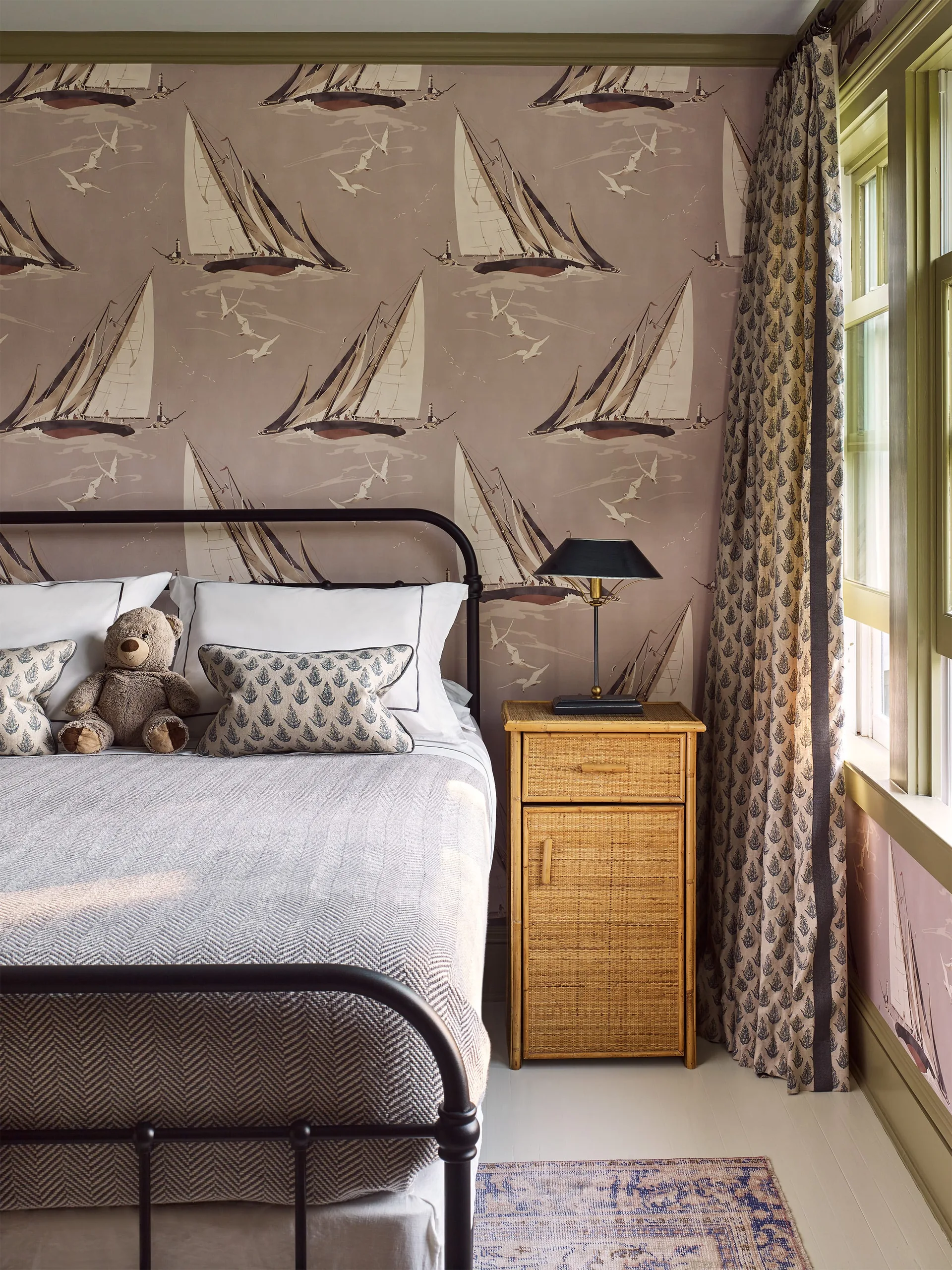 A cozy bedroom featuring sailboat-patterned wallpaper, a black metal bed frame, a rattan nightstand, and window trim painted olive green.