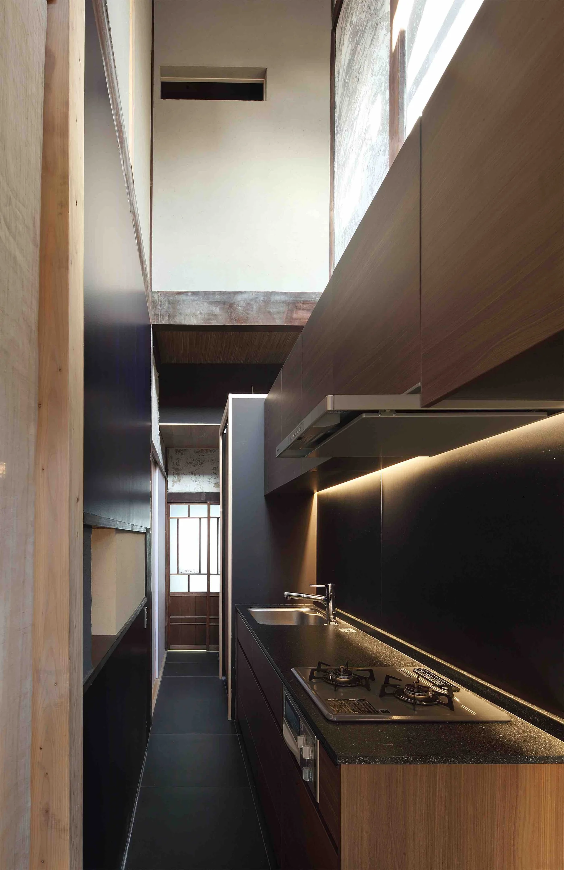A narrow modern galley kitchen features dark wood cabinets, a black countertop with a built-in stovetop, undercabinet lighting, and a double-height ceiling.