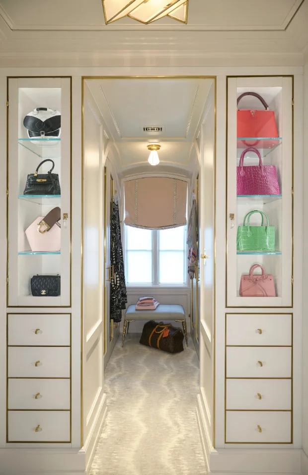 A symmetrical walk-in closet features white glass-front cabinets with gold trim displaying colorful handbags alongside a central window hallway.