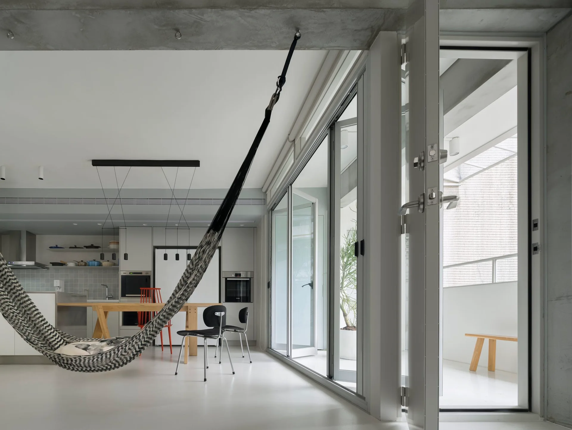 A minimalist open-plan living room features a striped hammock suspended from a concrete ceiling beam next to sliding glass doors that open to a balcony.