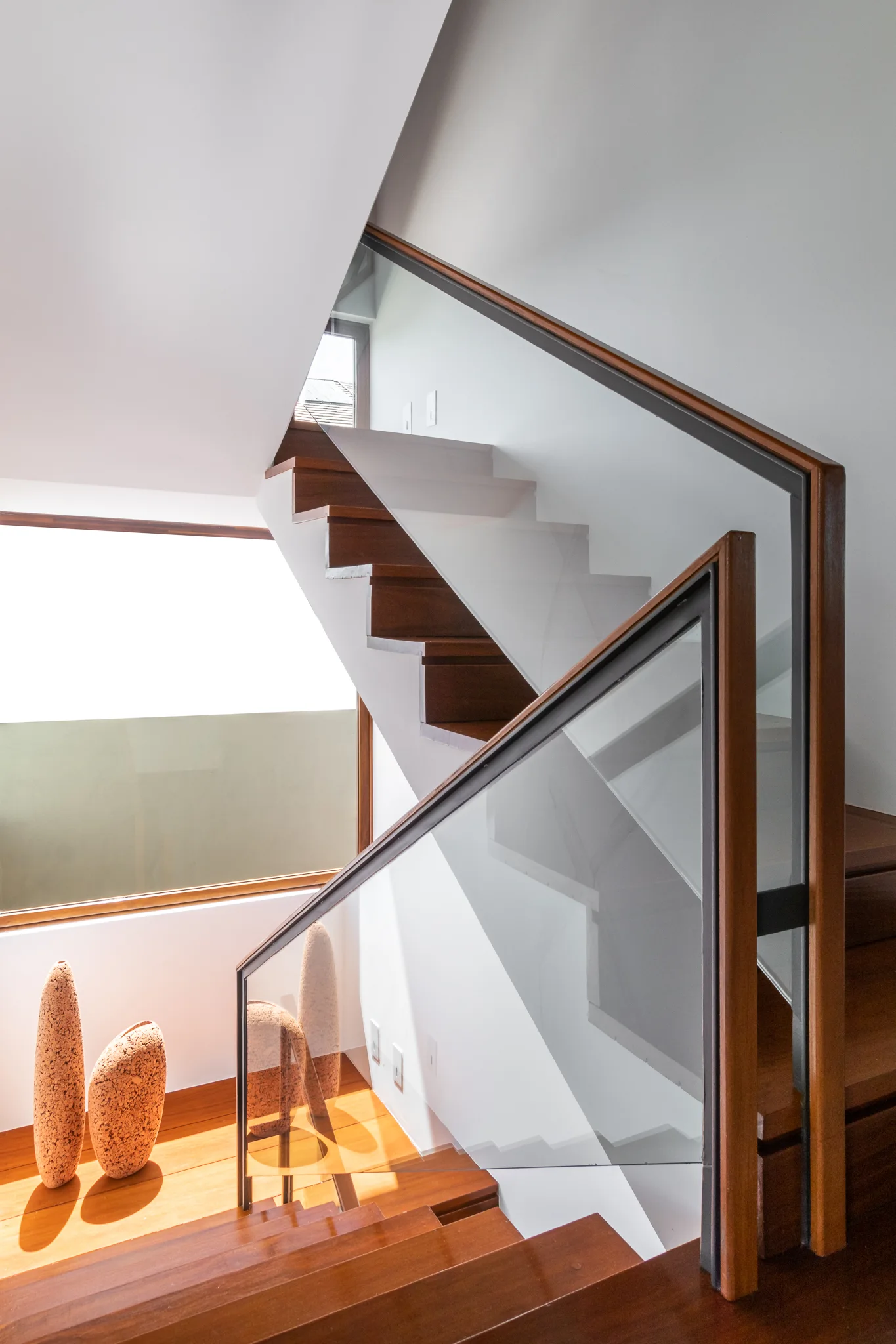 A modern floating staircase with rich wood treads and transparent glass railings stands next to a large window in a bright white hallway.