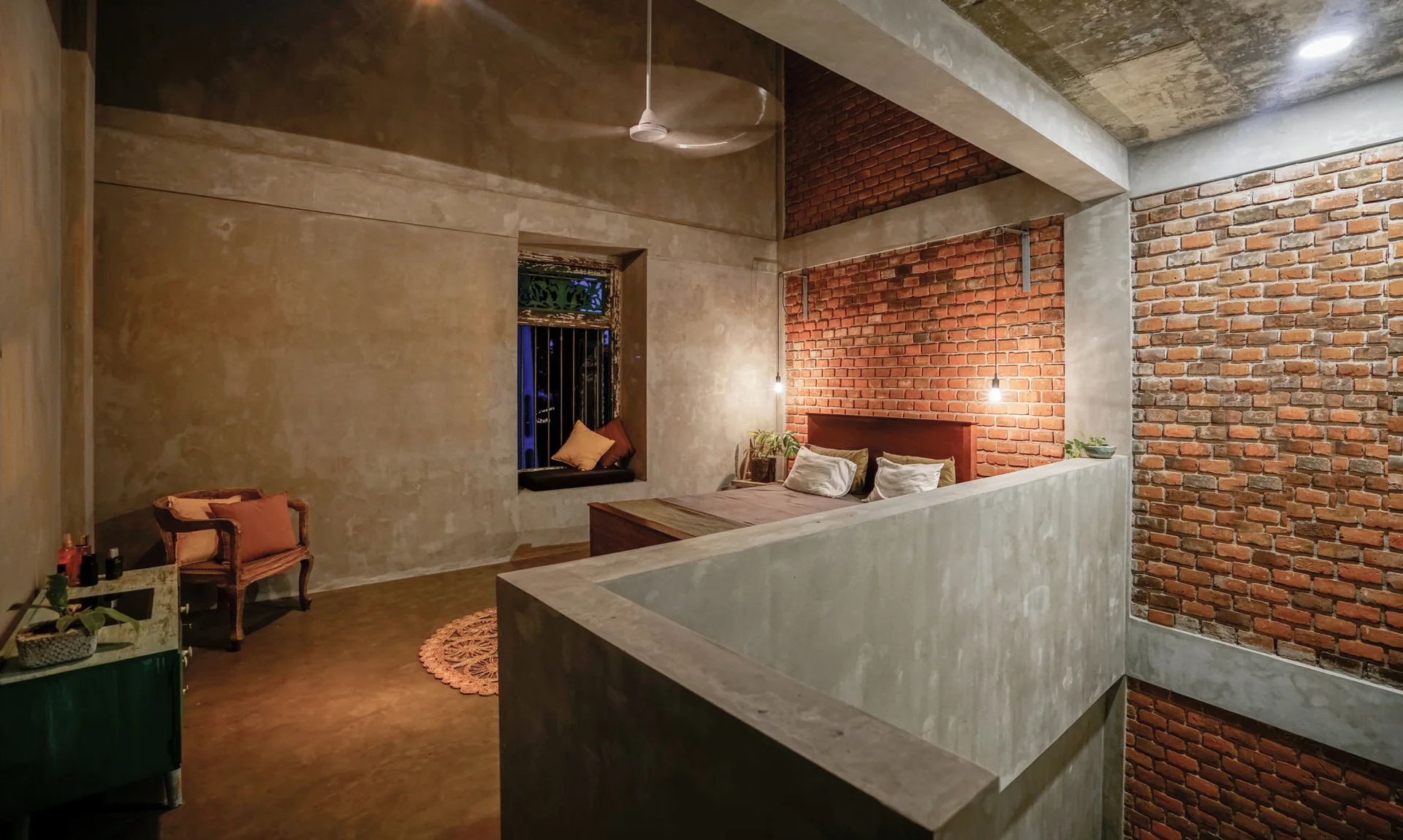 An industrial bedroom features an exposed red brick wall, smooth concrete surfaces, a wooden bed, and bare bulb pendant lights.
