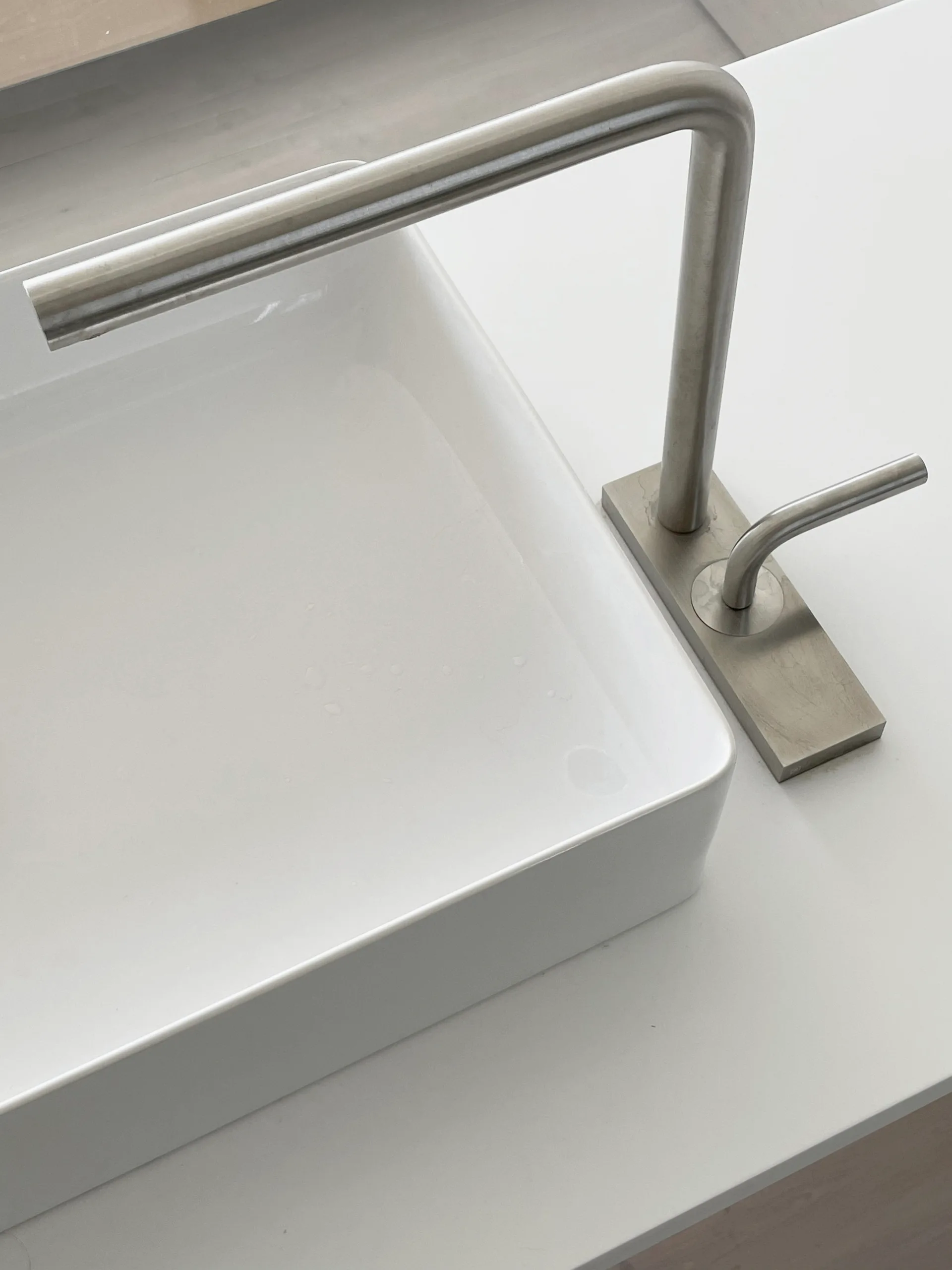 A close-up of a modern brushed metal faucet with a single lever handle next to a white rectangular ceramic vessel sink on a white countertop.