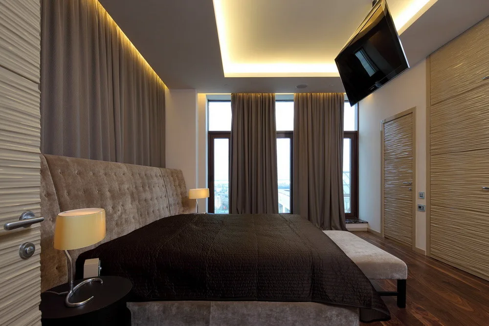 A contemporary bedroom featuring a large tufted headboard, dark bedding, wavy textured closet doors, and warm recessed cove lighting on the ceiling.