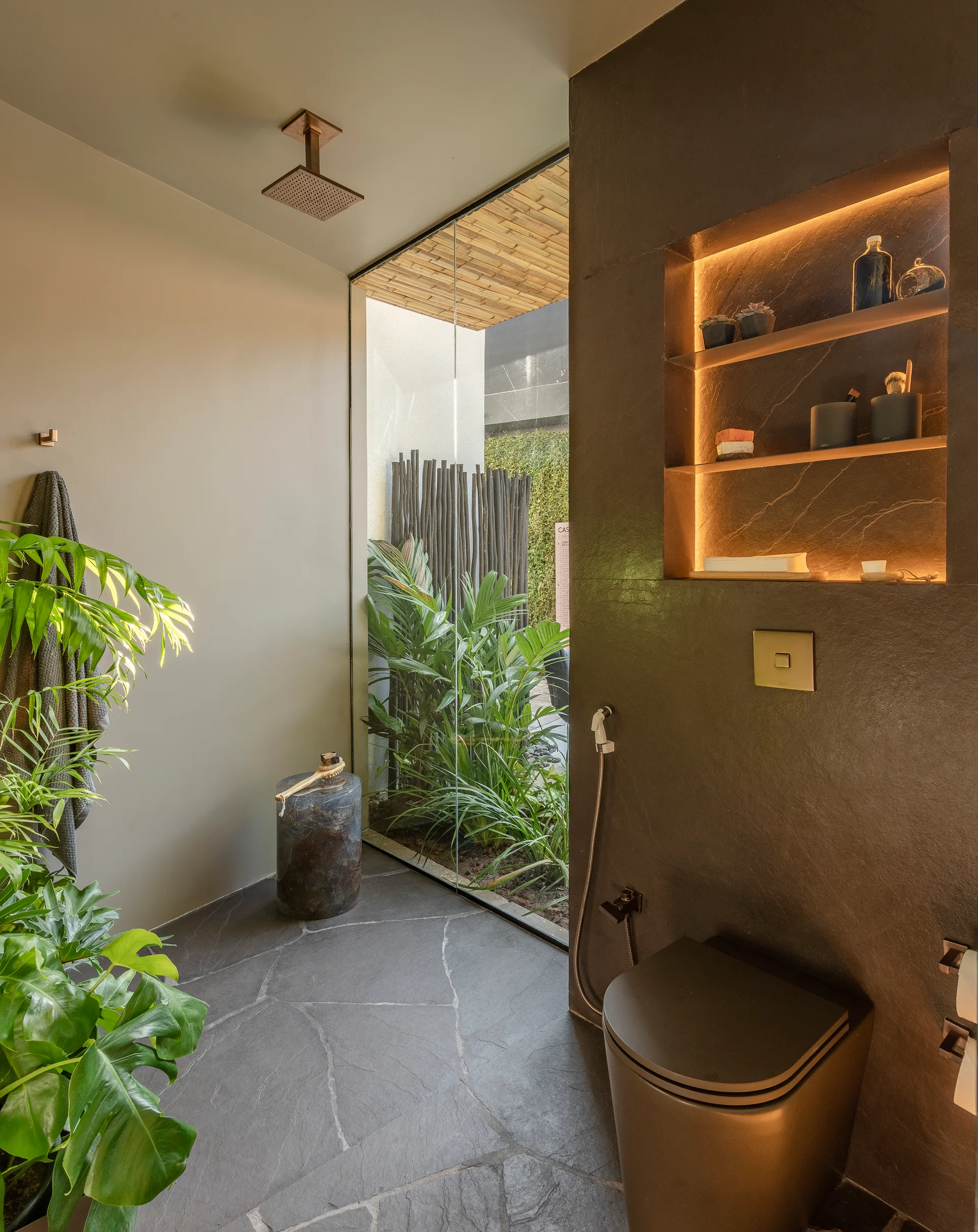 A moody dark grey bathroom featuring a black toilet, an illuminated recessed shelf niche, and a large glass window showing a green courtyard garden.