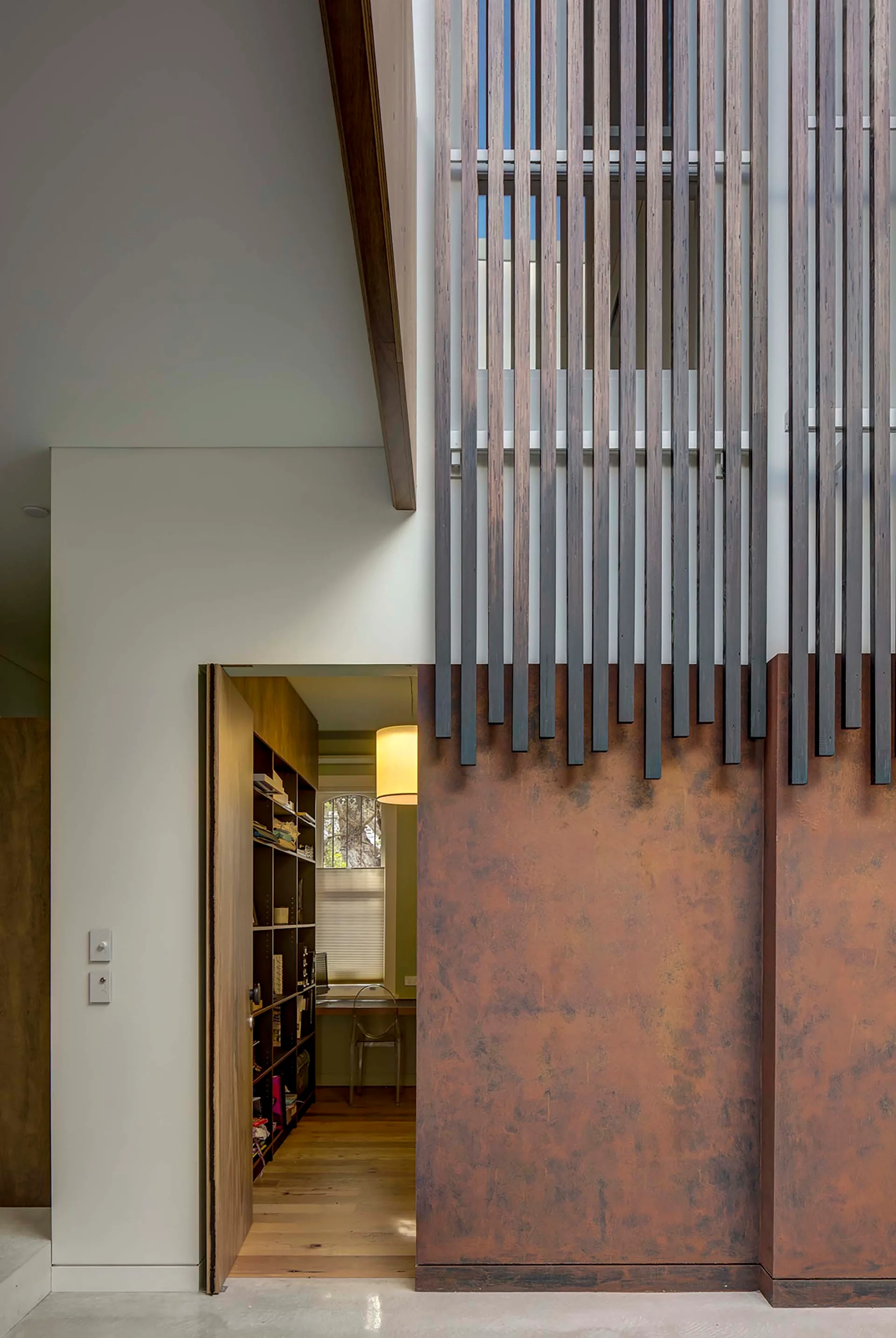An interior wall features dark vertical wooden slats over a rusted metal panel next to an open doorway leading into a study with bookshelves.