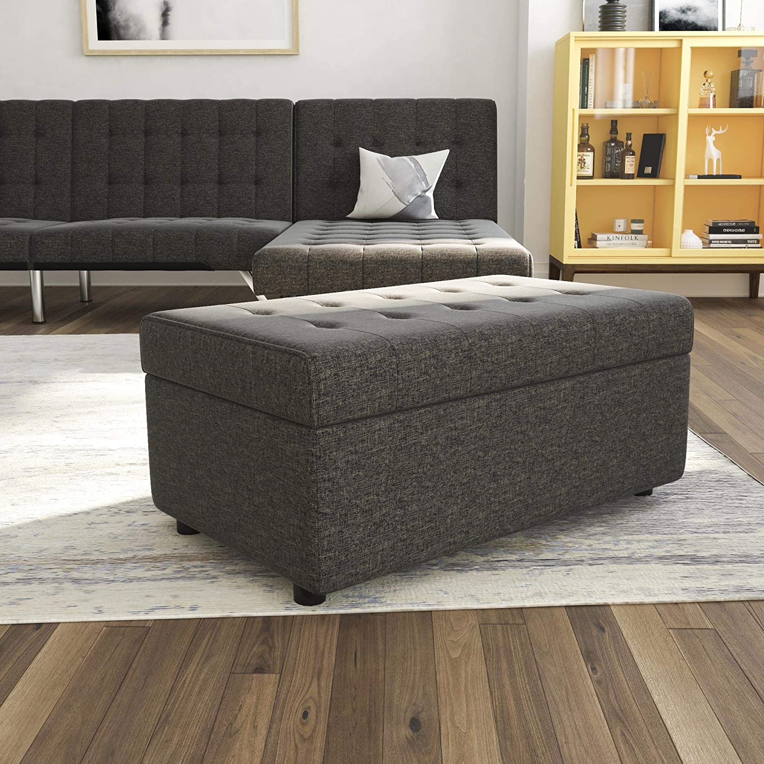 A dark grey tufted ottoman sits on a light area rug next to a matching sofa, with a bright yellow glass-front display cabinet in the background.