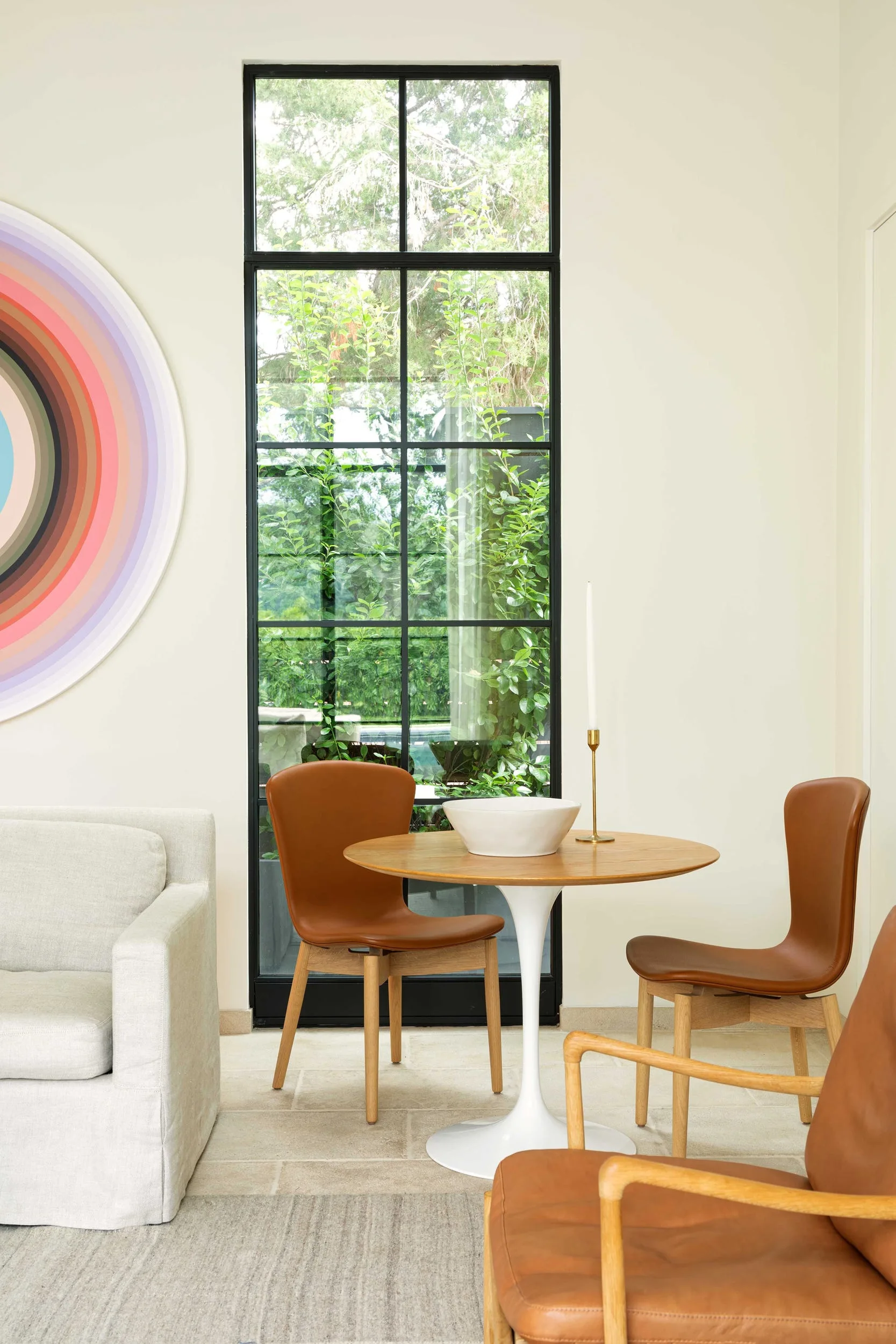 A small round wooden dining table and two brown leather chairs sit in front of a tall, black metal-framed window.