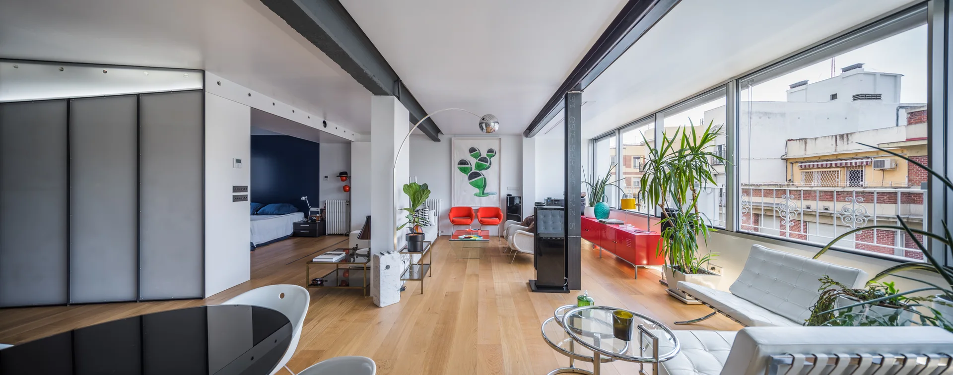 A spacious industrial apartment living room with light wood floors, exposed steel beams, a long red metal sideboard, and a wall of windows.