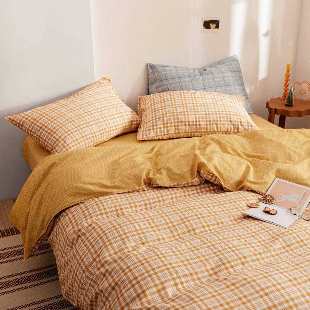A cozy bed is dressed in yellow checkered sheets and a polka-dot duvet cover, with a grey accent pillow resting against a plain white wall.
