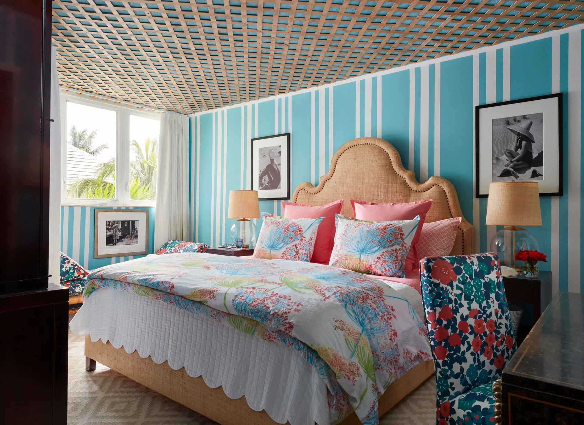 A coastal bedroom features a bed with a tan headboard, vibrant floral bedding, blue and white striped wallpaper, and a wooden lattice ceiling.