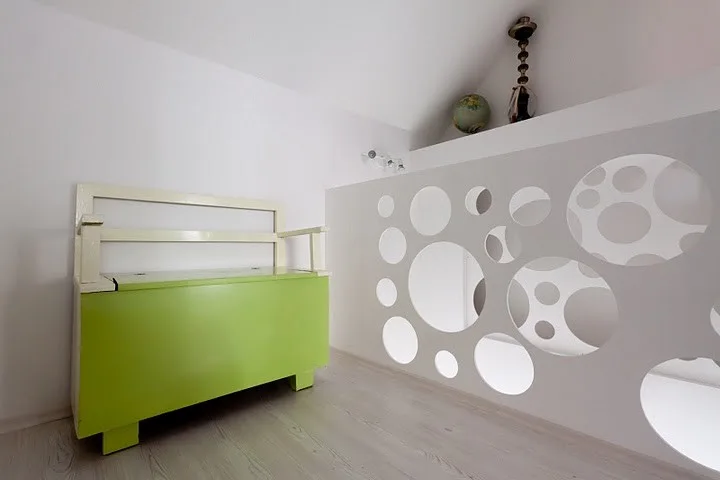 A light-filled loft space features a lime green storage bench next to a white divider wall with circular cutouts under a sloped ceiling.