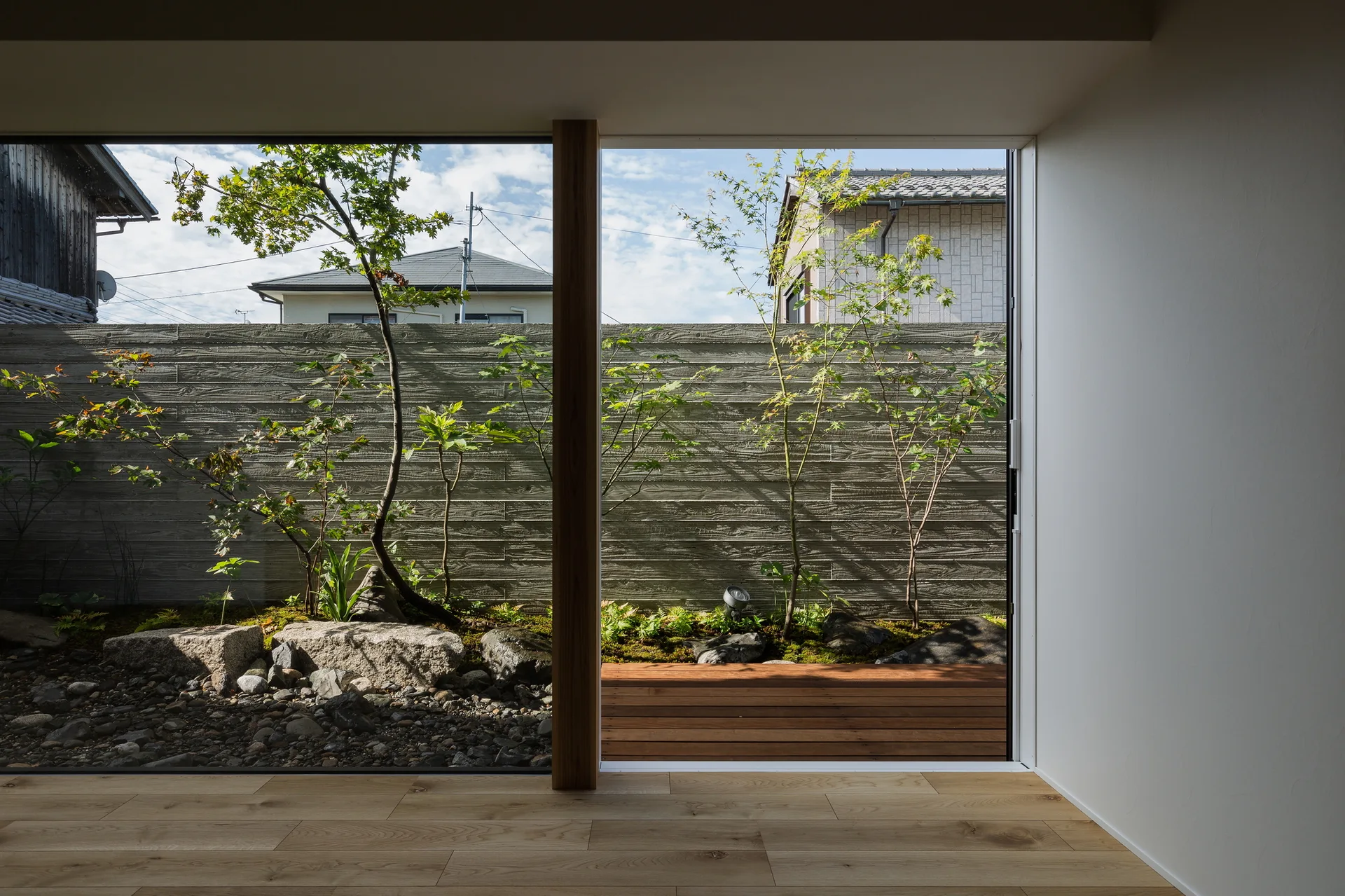 A minimalist room features light wood floors and large windows opening onto a quiet courtyard garden with a textured concrete wall and small trees.
