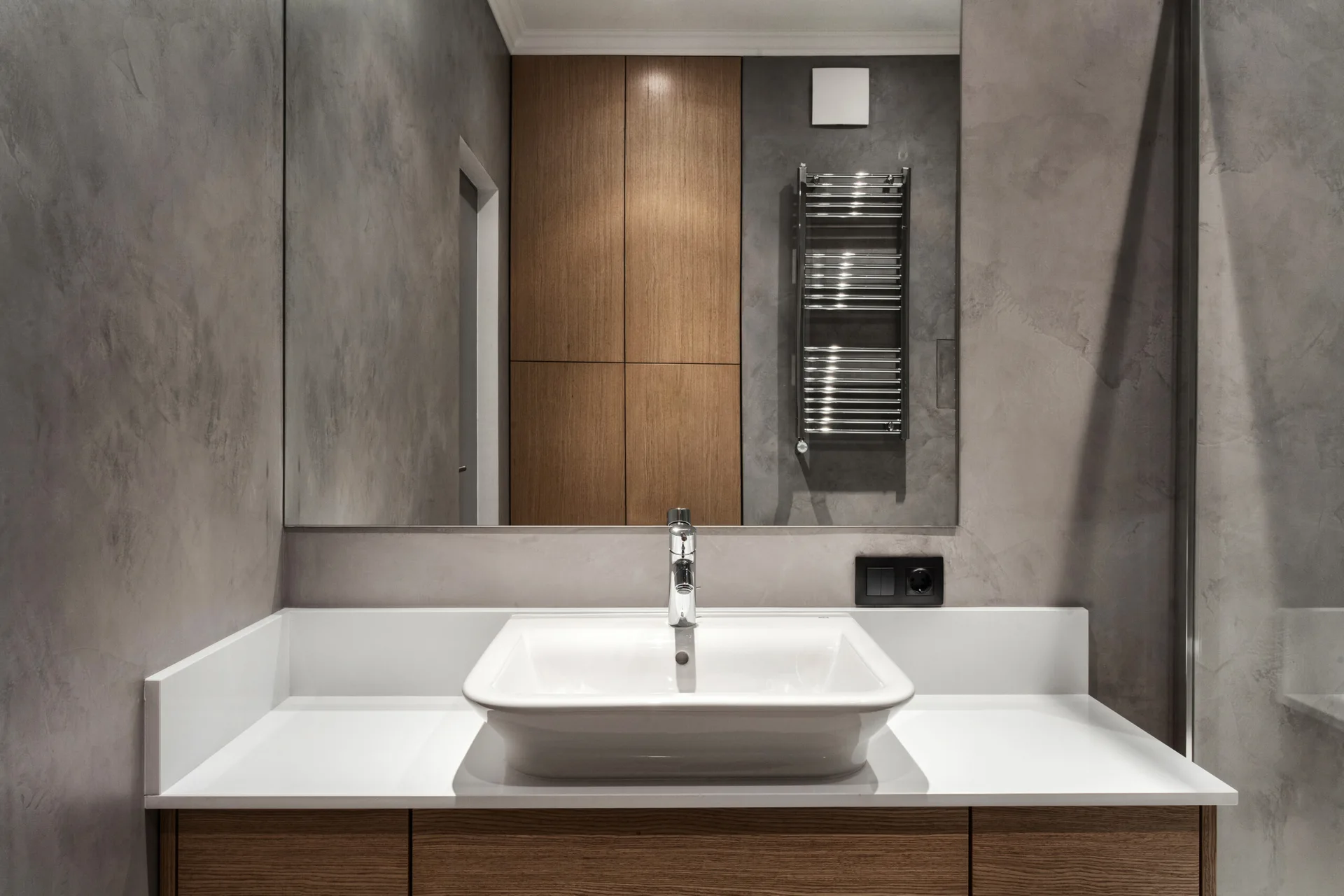 A modern bathroom vanity featuring a white sink on a wood cabinet set against textured grey plaster walls under a large frameless mirror.