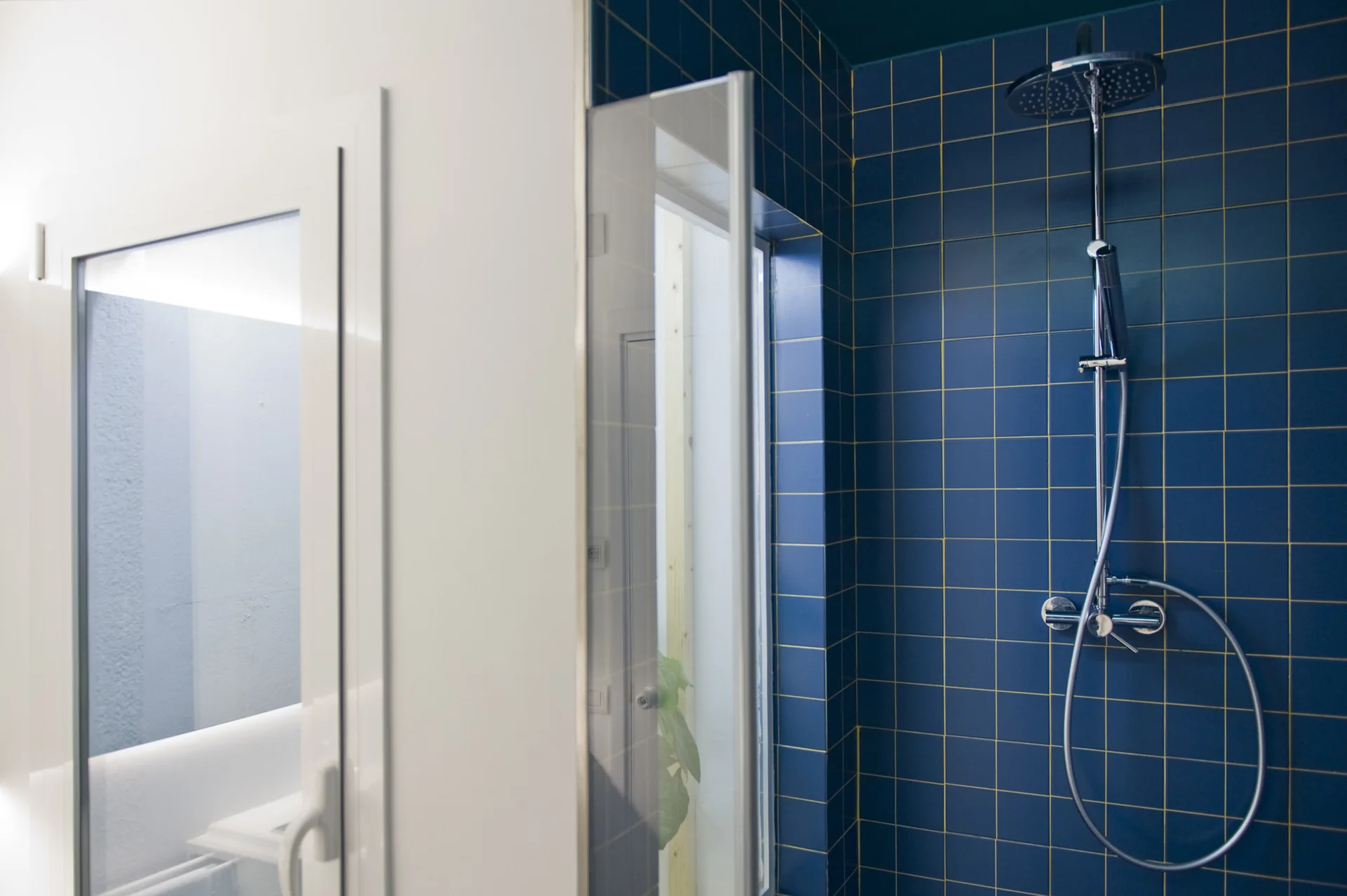A modern shower enclosure features glossy blue square tiles with contrasting gold grout, a chrome shower head, and a glass door.