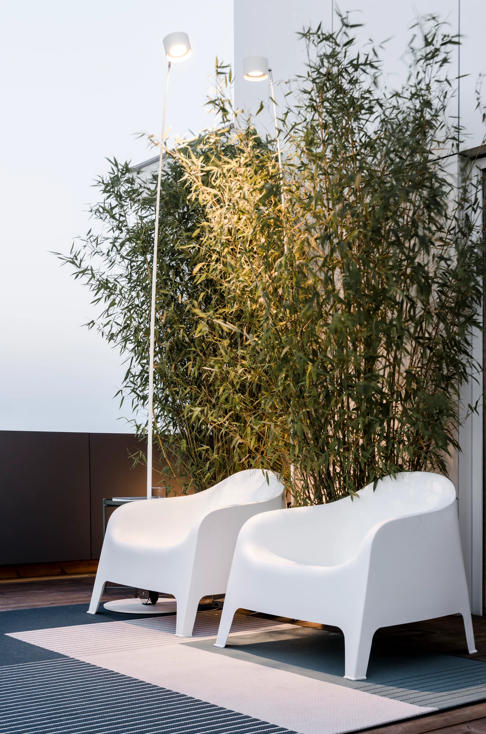Two white molded plastic armchairs sit on a striped rug on a wooden terrace next to tall bamboo plants and slim floor lamps.