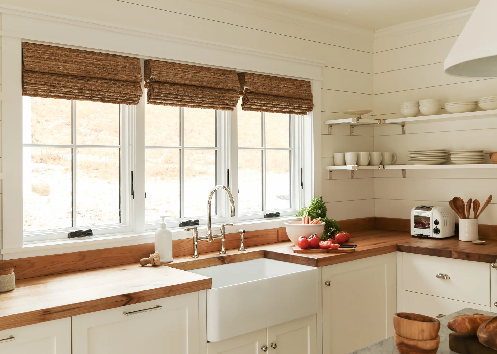 A farmhouse kitchen featuring a white apron sink, natural wood countertops, and woven shades on a triple window.