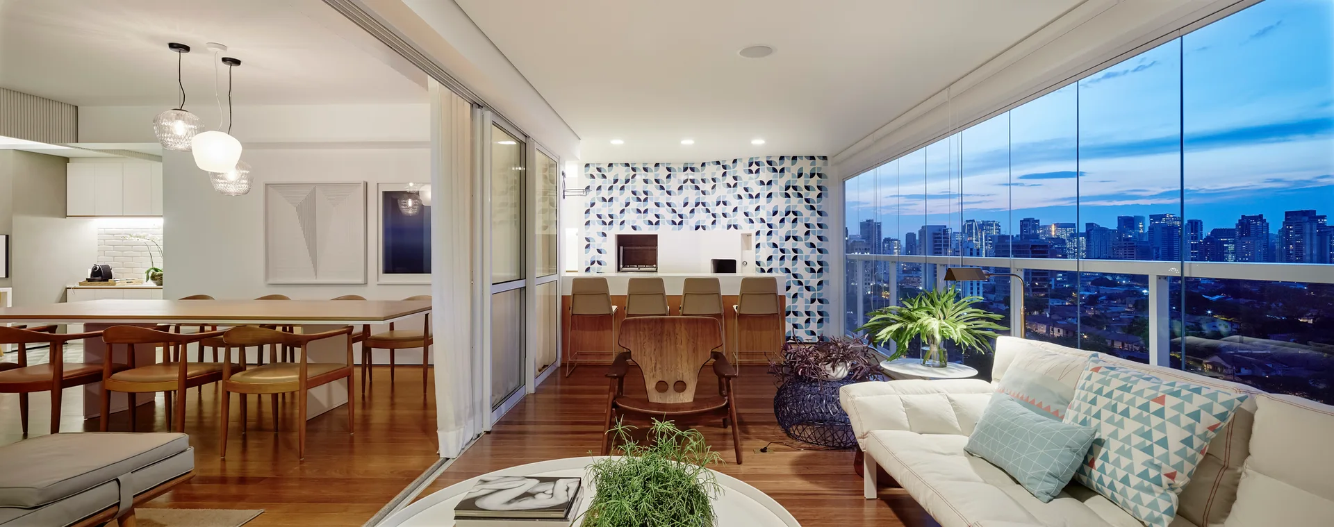A modern open-concept apartment interior features a dining area, a living area with a sofa, and a glass-enclosed balcony with a blue geometric tile wall.